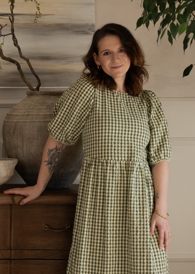 Vrouw in groene geruite jurk leunt op houten ladekast met grote vaas, gezellige binnenruimte.