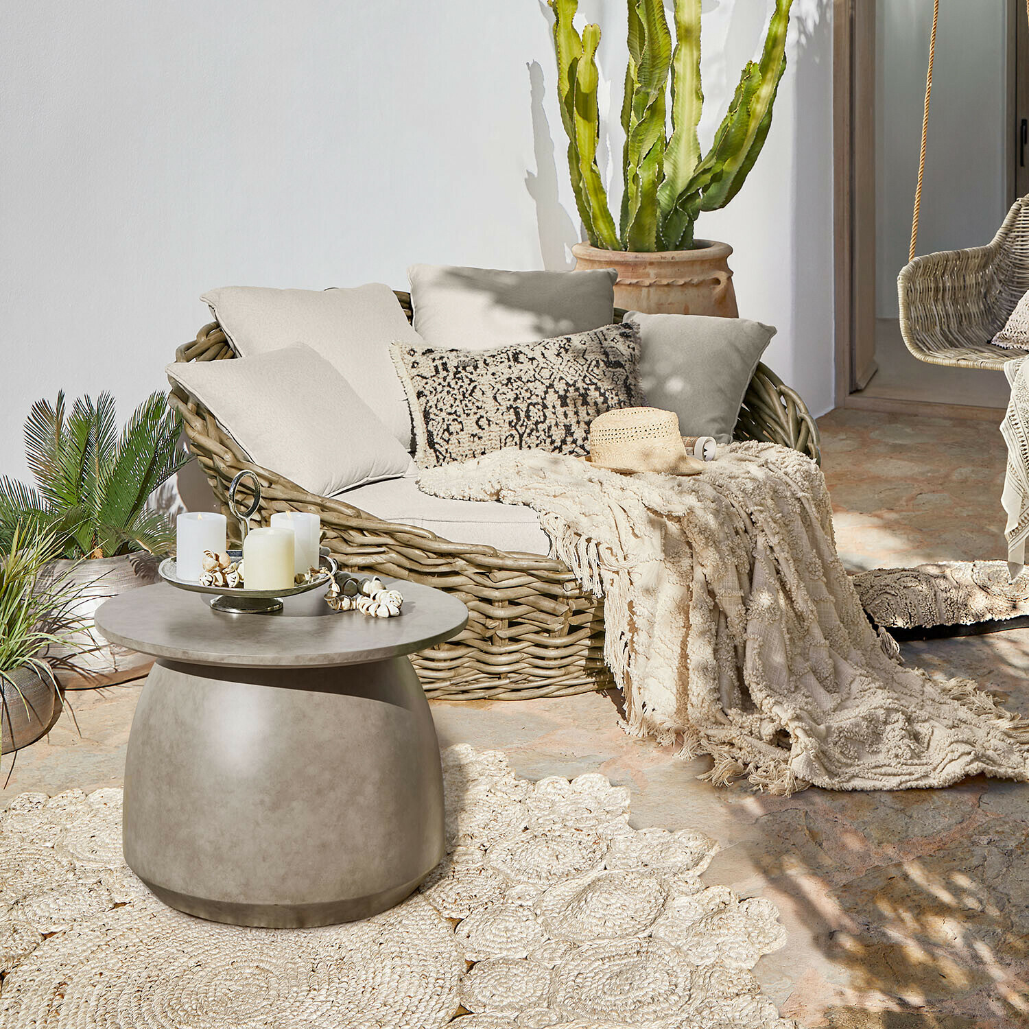 Ronde rotan bank met crèmekleurige kussens en deken op terras met betonnen salontafel