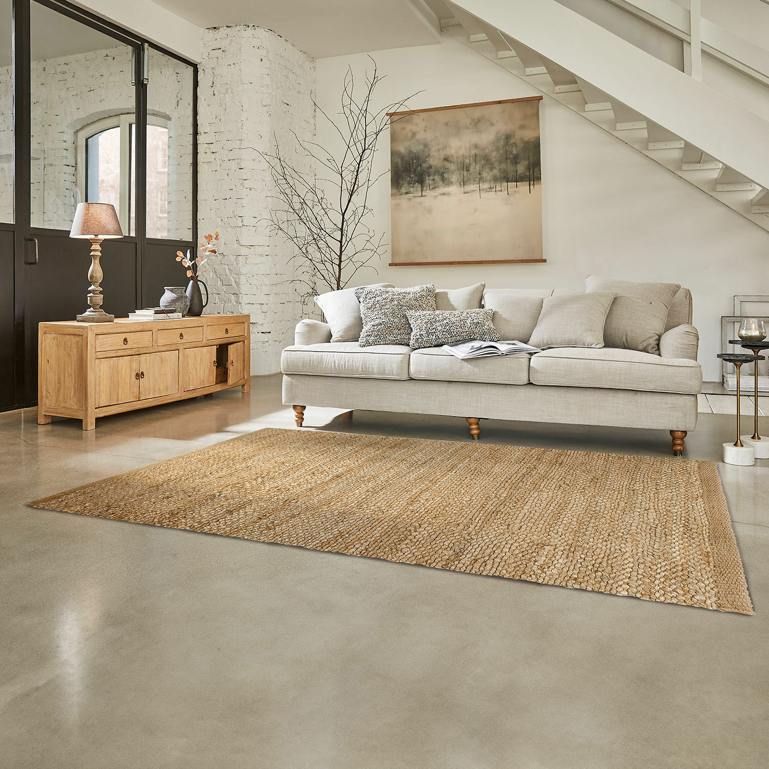 Beige bank met kussens en lichte houten dressoir in lichte woonkamer met natuurlijke jute vloerkleed