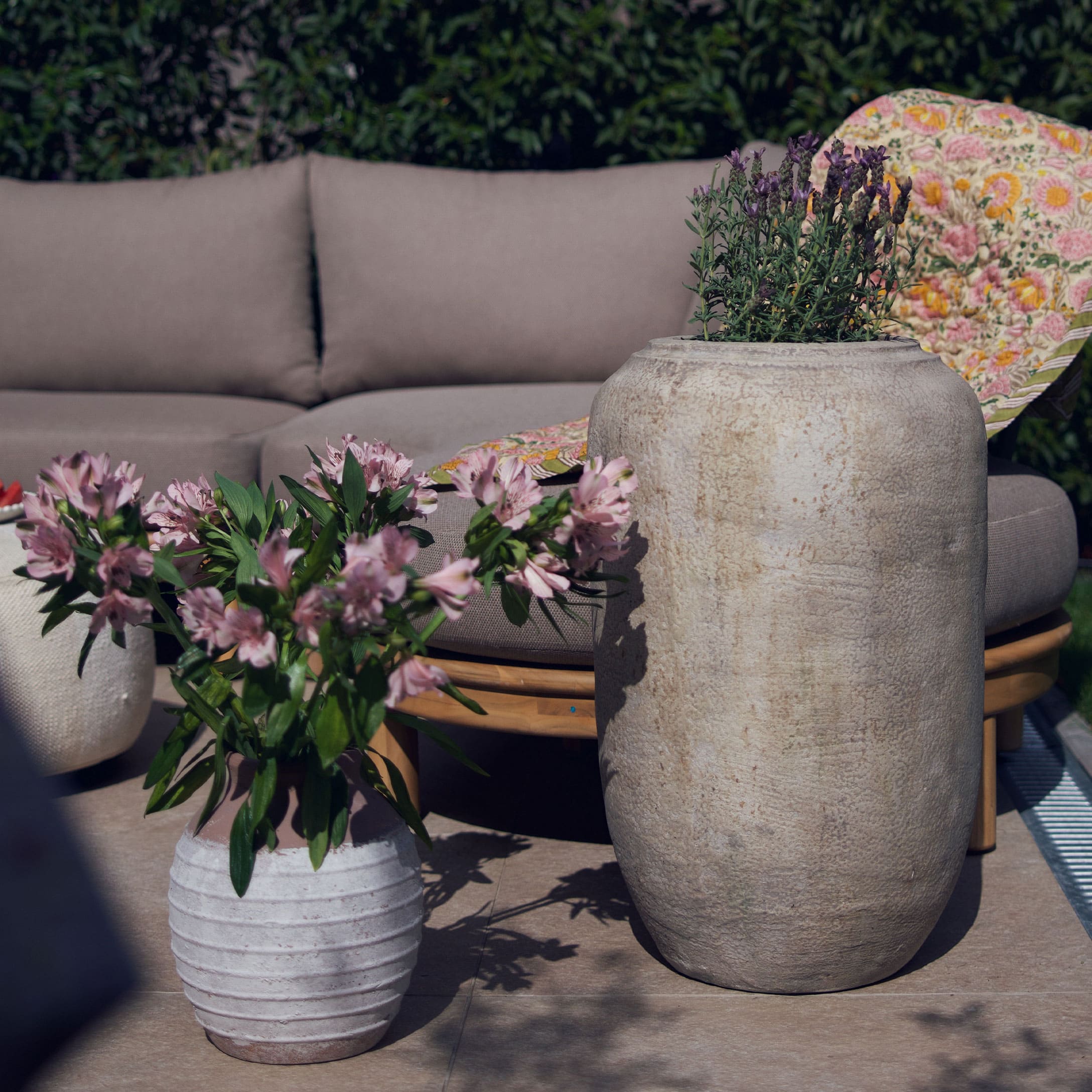 Grote en kleine lichte stenen pot met bloemen op terras met grijze bank en kleurrijk deken