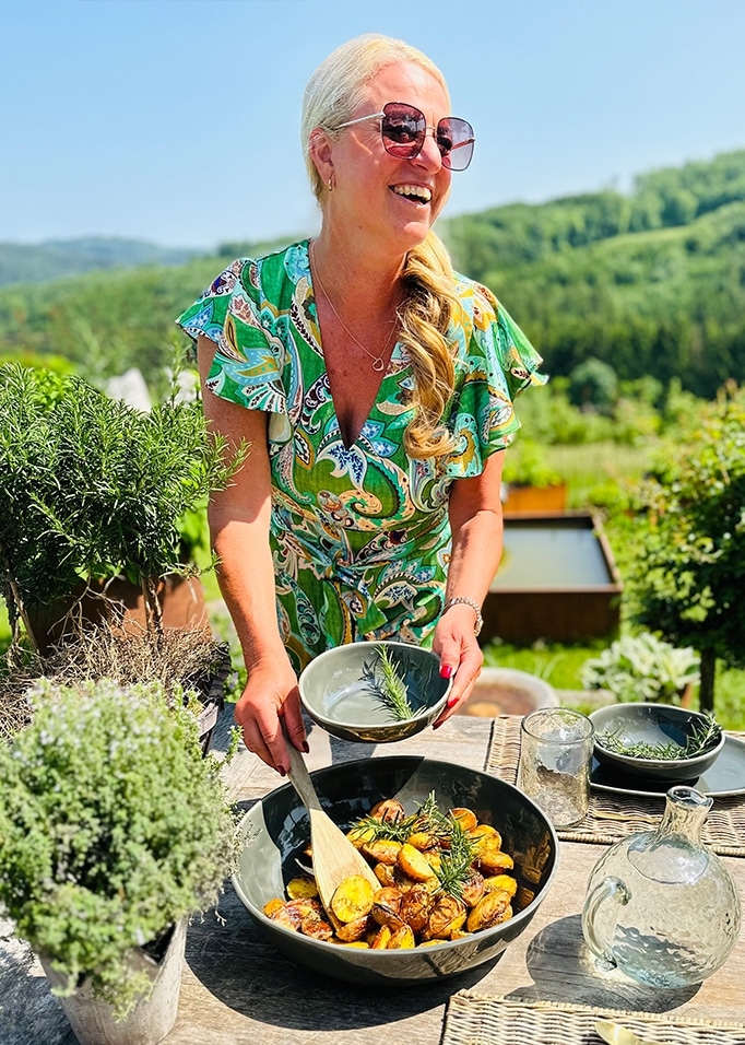Vrouw in groene jurk serveert gebakken aardappelen in keramische kom buiten met kruiden en natuur