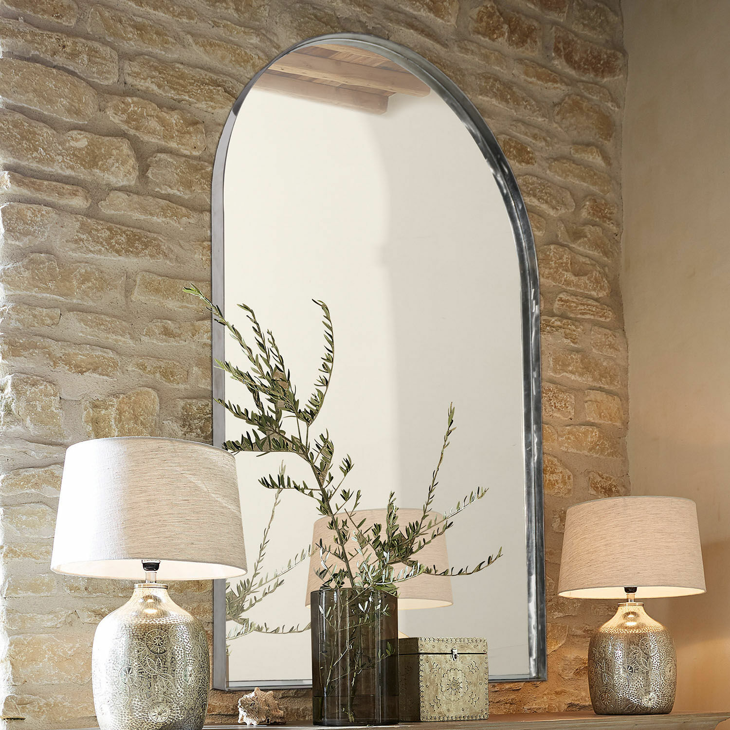 Grote spiegel met metalen lijst tegen stenen muur, verlicht door lampen op houten plank met decoraties