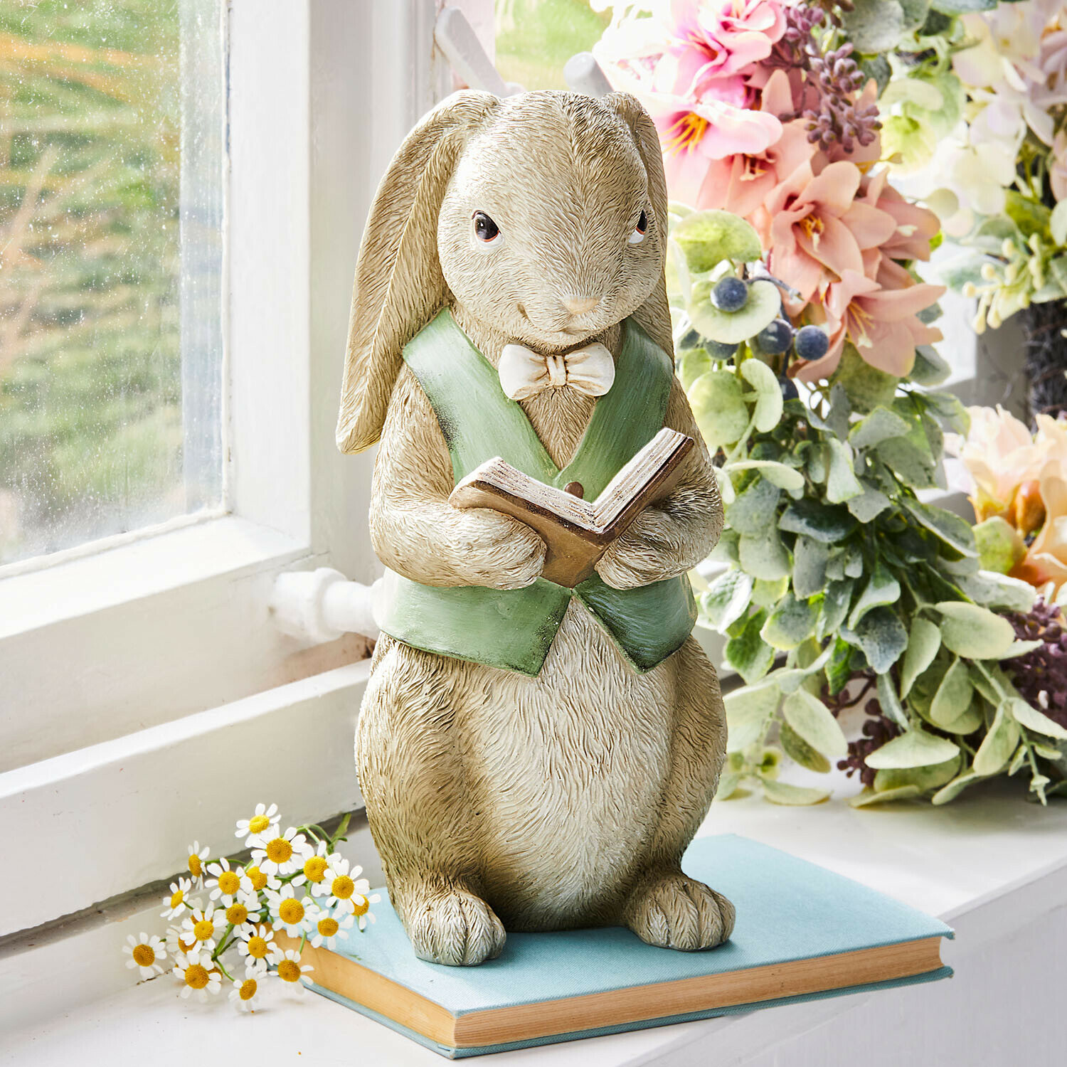 Konijnenbeeld van hars met groene vest lezend boek op vensterbank met bloemen