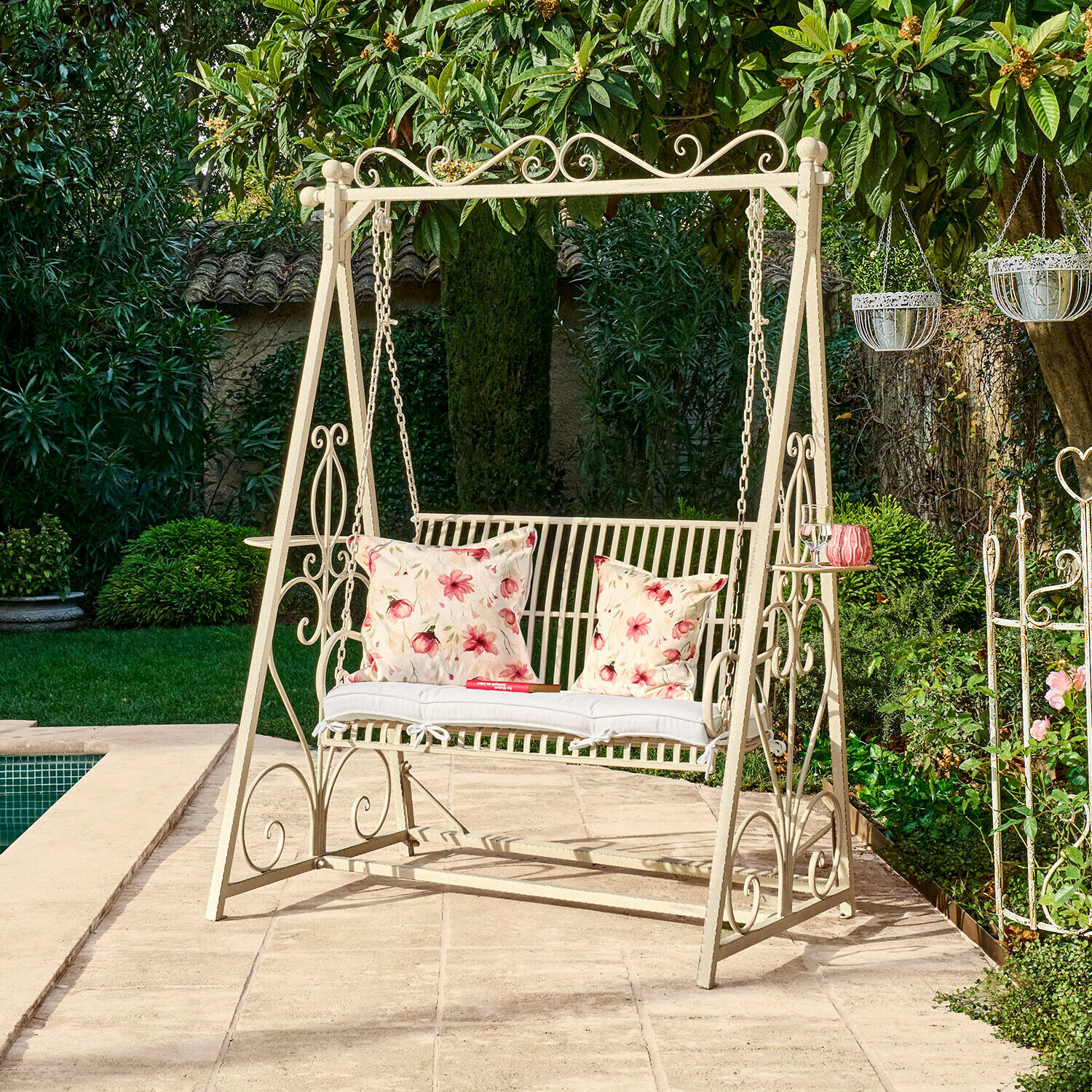 Metalen tuinschommel in crèmekleur met bloemenkussens op terras bij zwembad