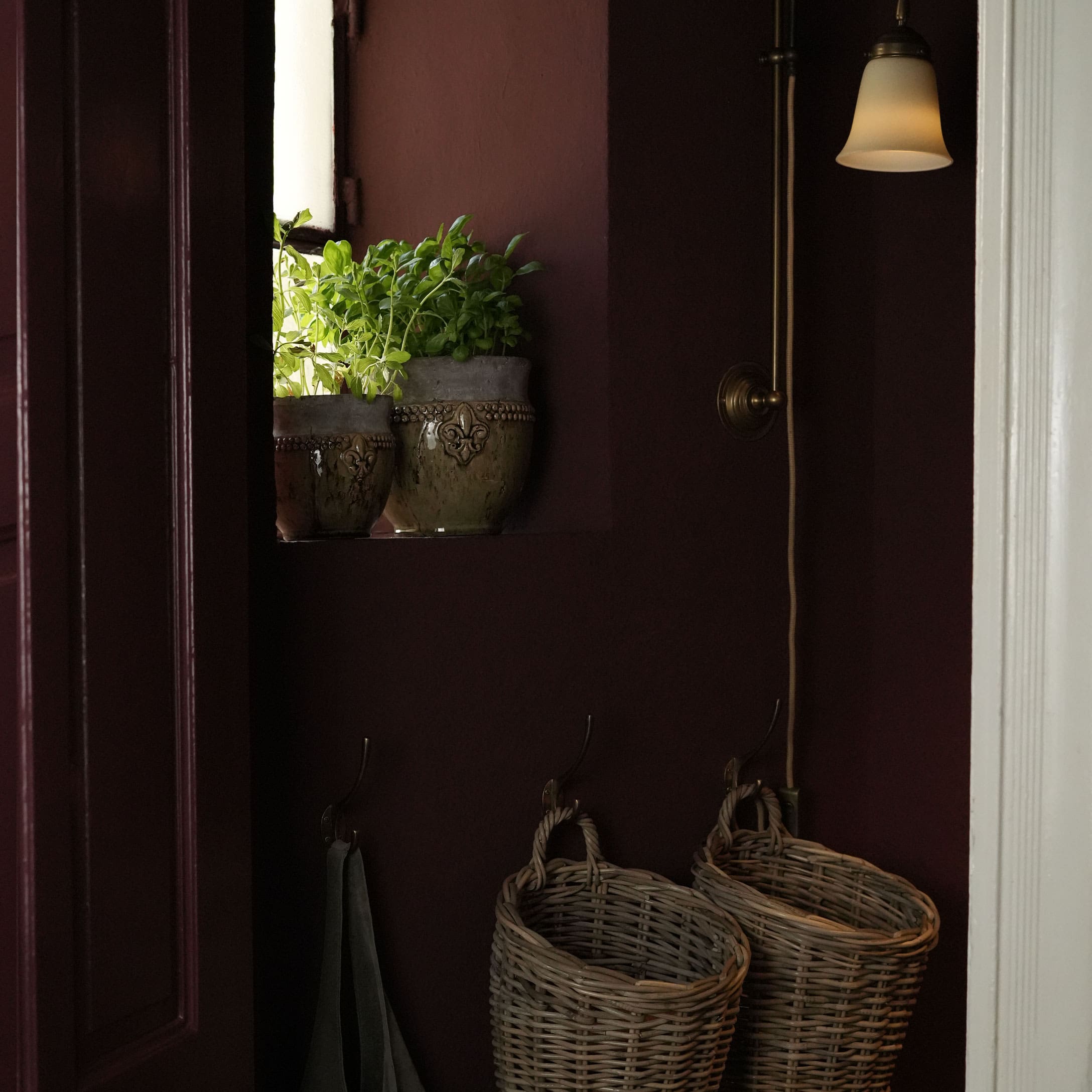 Twee keramische bloempotten met groene planten tegen een bordeauxrode muur en een wandlamp met matglas kap