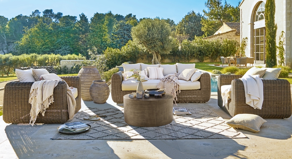 Bruin rieten tuinmeubelset met crèmekleurige kussens en ronde salontafel op zonnig terras