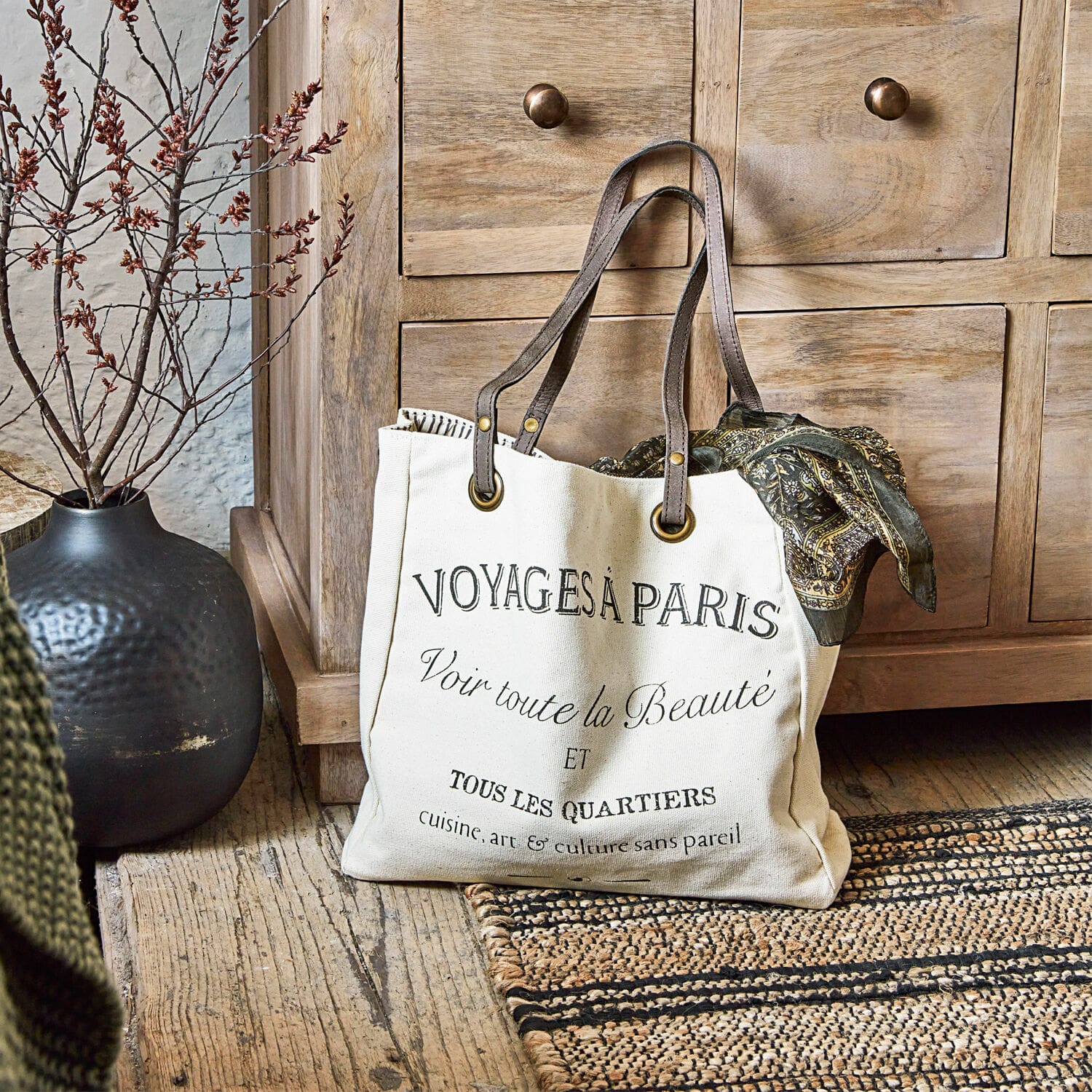 Tas Voyage Beige katoenen tas met zwarte opdruk en bruine handvatten op houten vloer