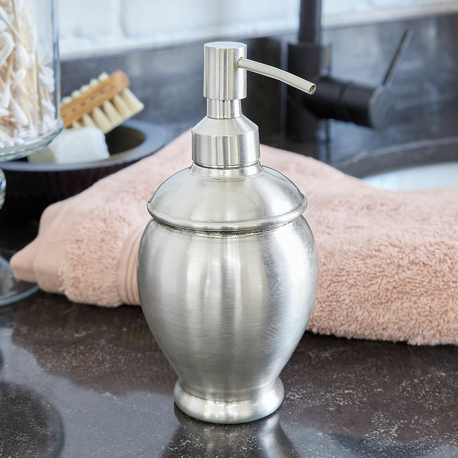 Zilverkleurige metalen zeepdispenser op wastafel met roze handdoek op de achtergrond