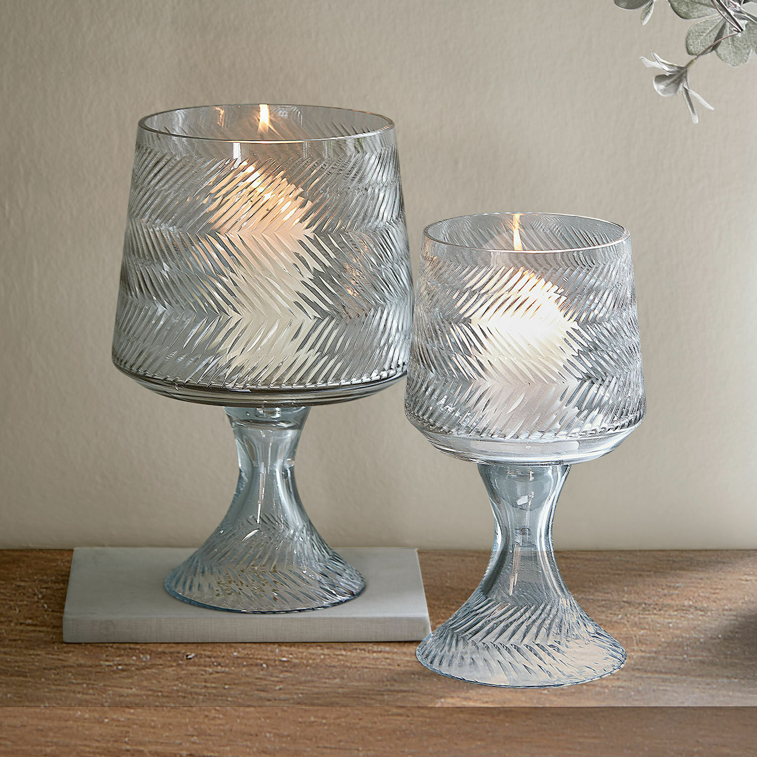 Twee transparante glazen windlichten met reliëfpatroon en witte kaarsen op houten tafel