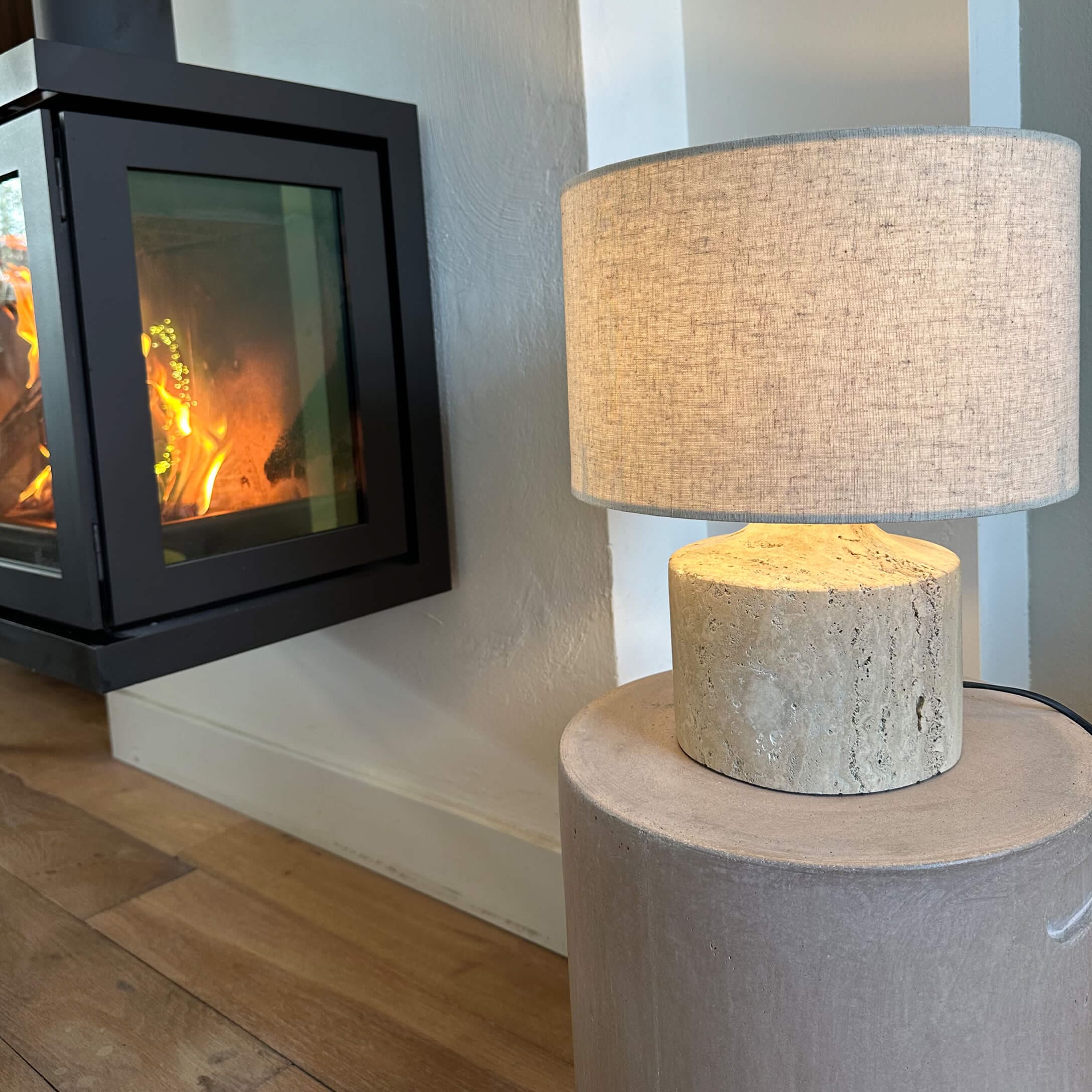 Tafellamp met beige kap en lichte stenen voet naast brandende open haard