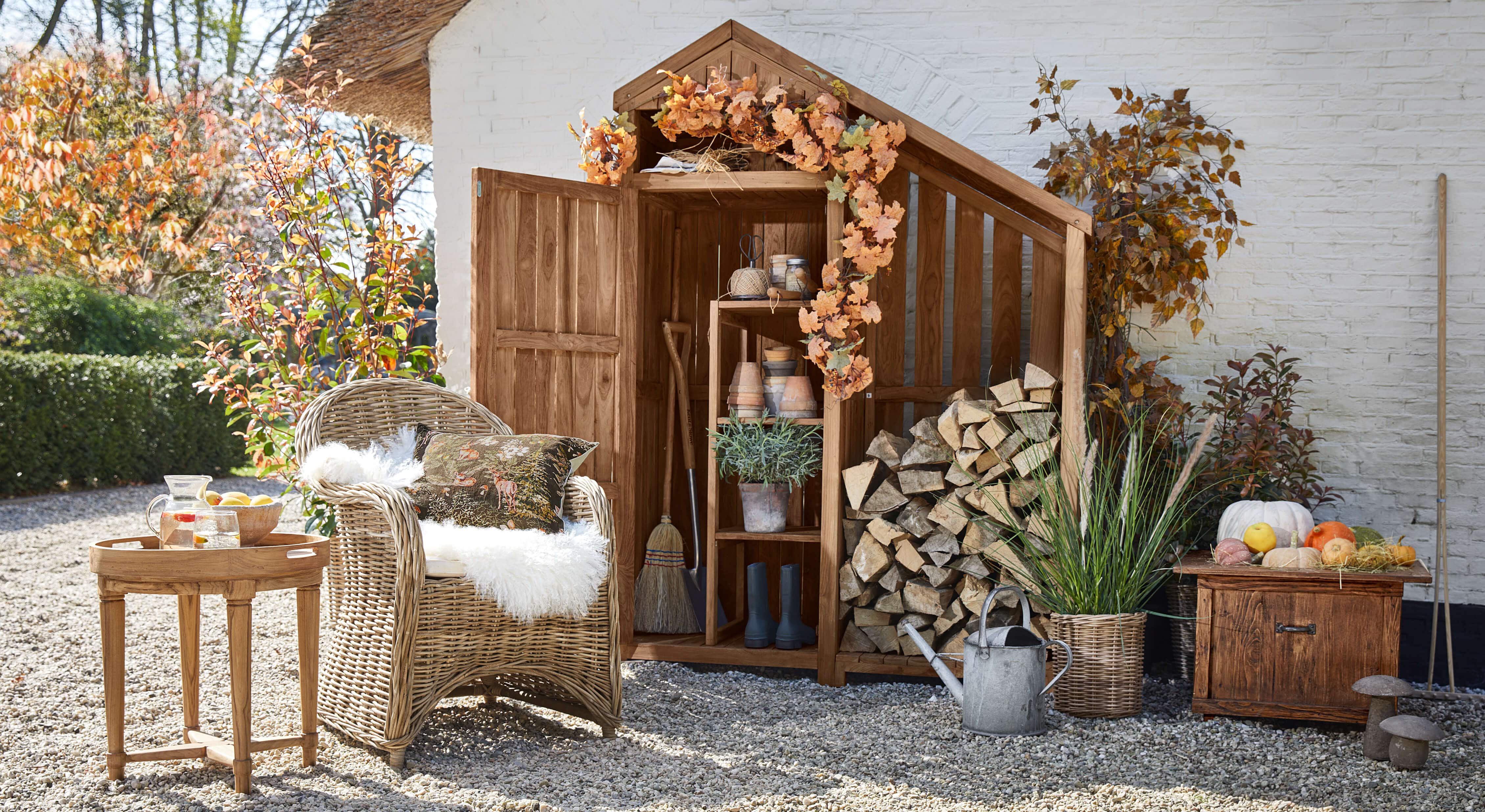Houten tuinhuisje met herfstdecoratie, rotan stoel en tafeltje met karaf op grindvloer