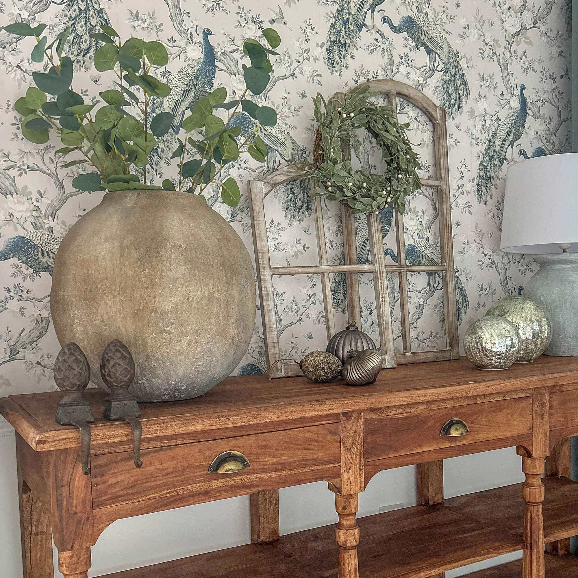 Houten tafel met grote aardewerken vaas, groene bladeren, lamp en decoraties erop