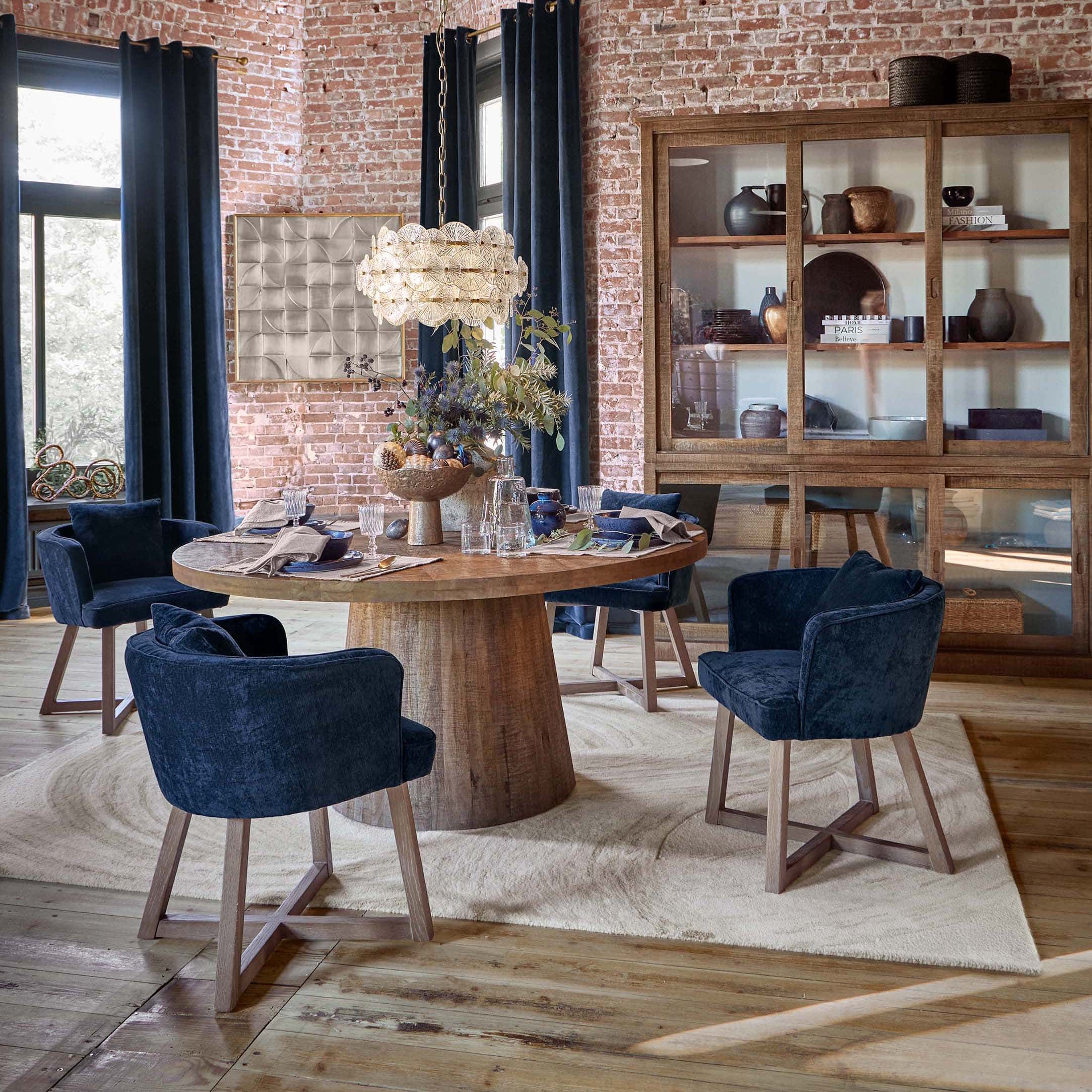 Eetkamer met ronde houten tafel en donkerblauwe fluwelen stoelen voor een bakstenen muur en houten vitrinekast