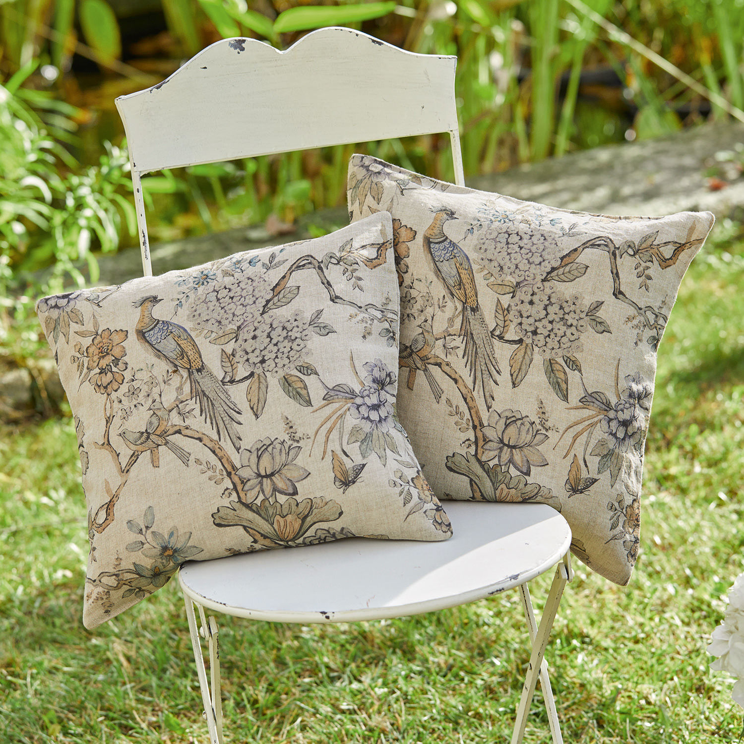 Kussenhoes set van 2 Lorry Twee decoratieve katoenen kussens in beige met vogel- en bloemenprint op een witte stoel in de tuin