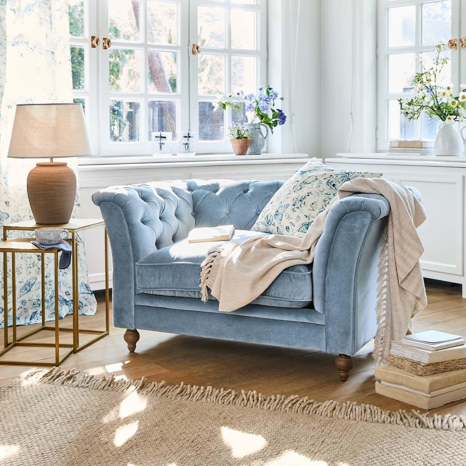 Blauwe fluwelen tweezitsbank met bloemkussen en beige deken in lichte woonkamer met houten vloer