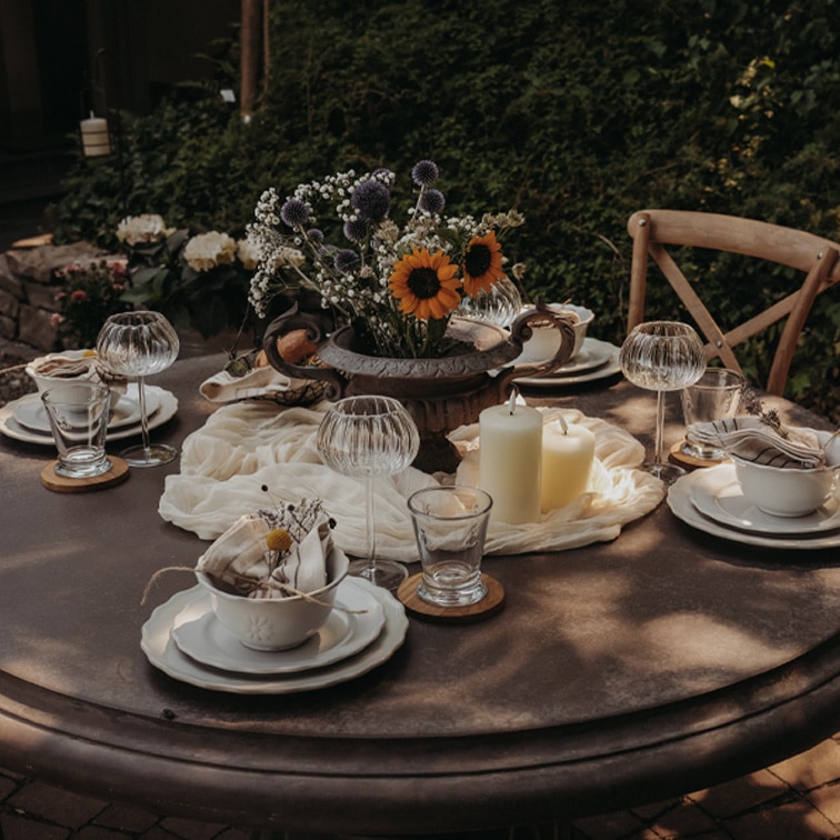 Ronde tuintafel met kaarsen en bloemen in metalen vaas, klaar voor een buitenbijeenkomst.