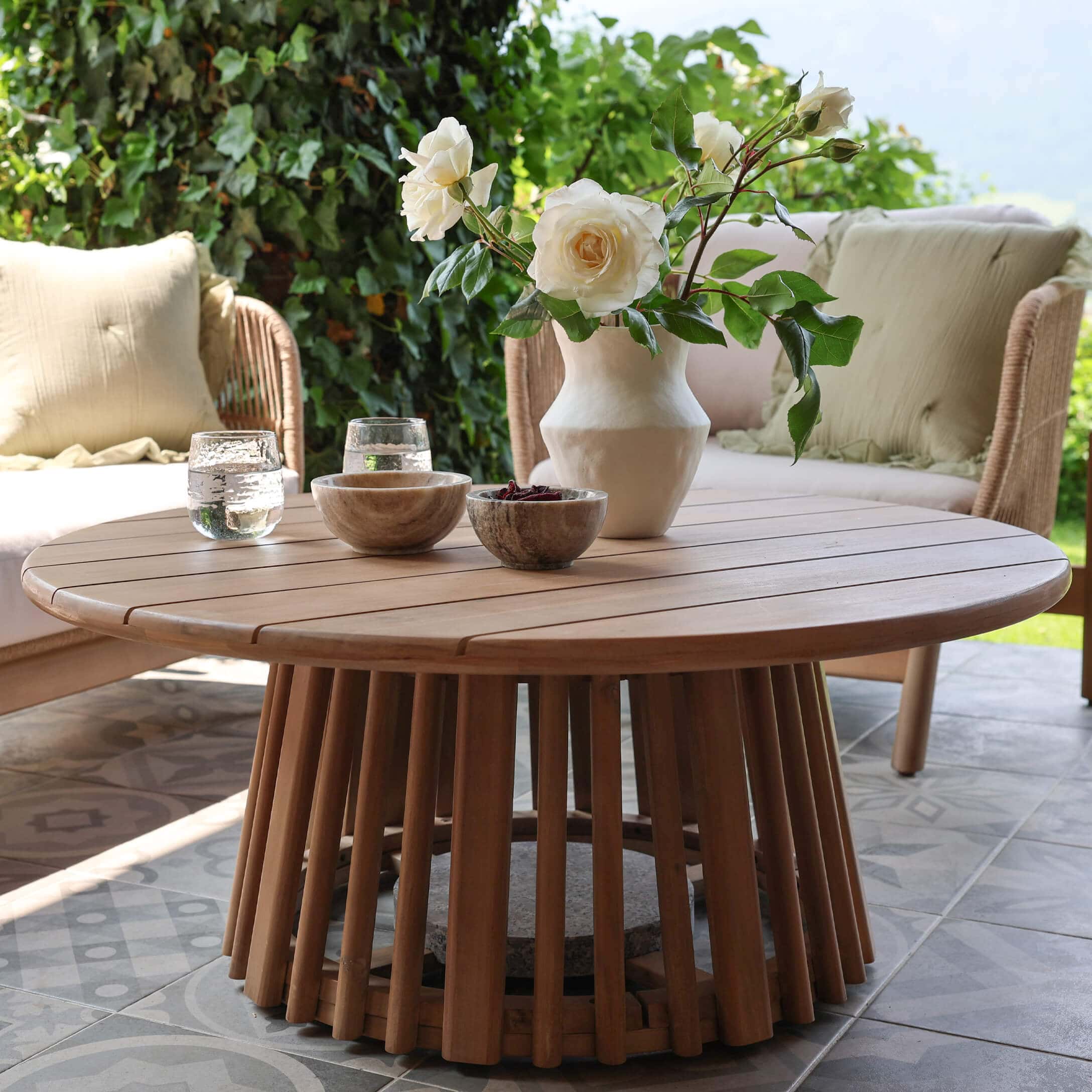 Ronde salontafel van natuurlijk teakhout op terras met bloemendecoratie en stenen kommen