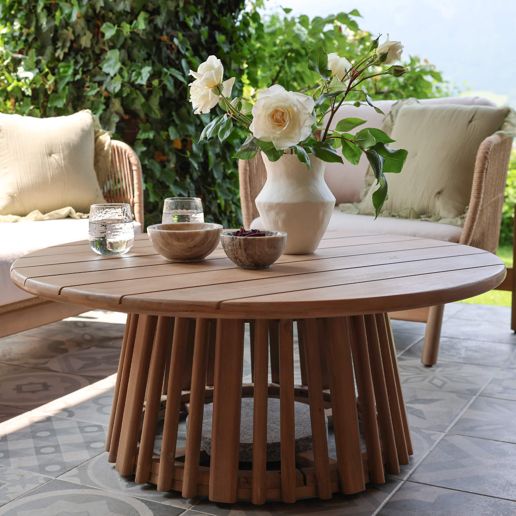 Ronde salontafel van natuurlijk teakhout op terras met bloemendecoratie en stenen kommen