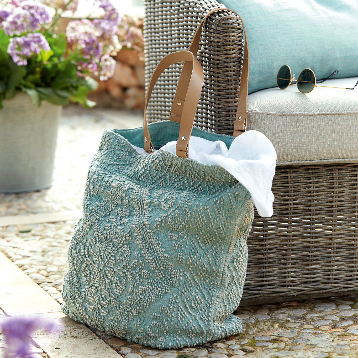 Turquoise tas van gestructureerde stof met bruine handvatten naast een rotan stoel en zonnebril