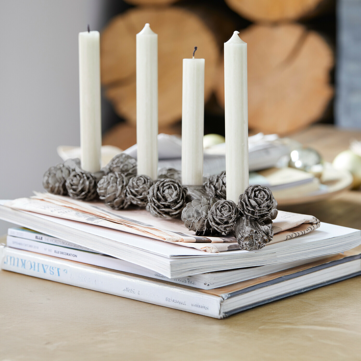Kaarsenhouder Arasta Decoratieve kandelaar met grijze dennenappelornamenten en vier witte kaarsen op een stapel tijdschriften