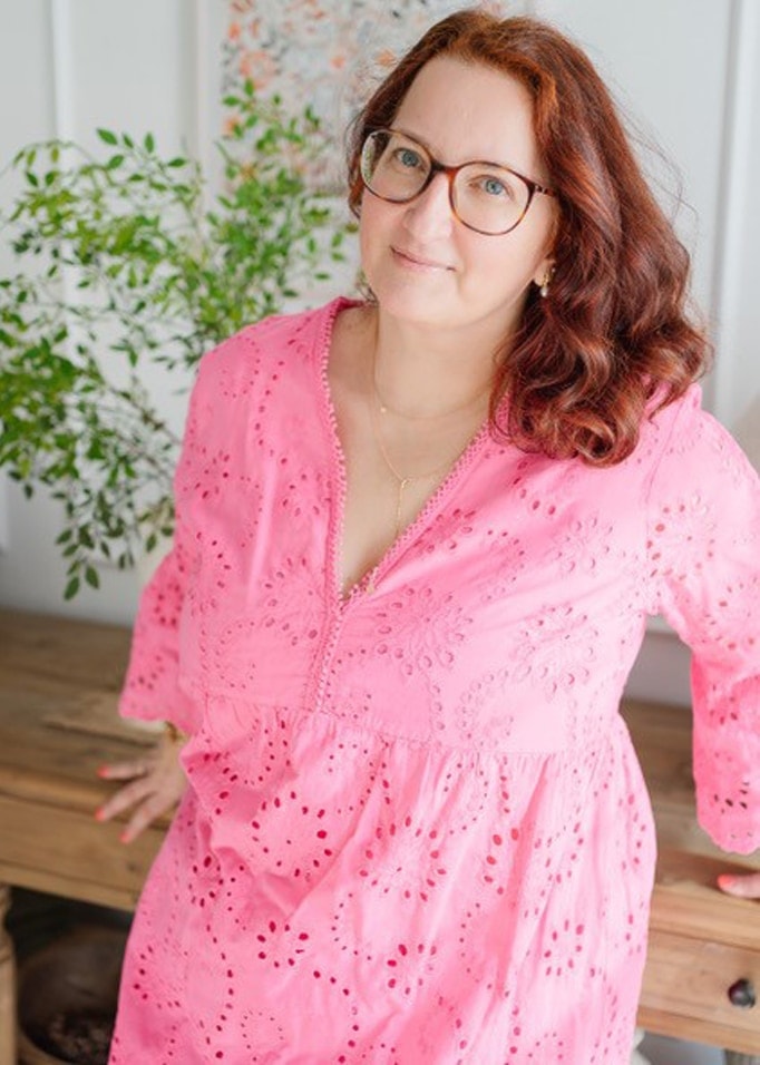 Vrouw in roze opengewerkt jurkje met fijn patroon, leunend op houten tafel met plant op de achtergrond
