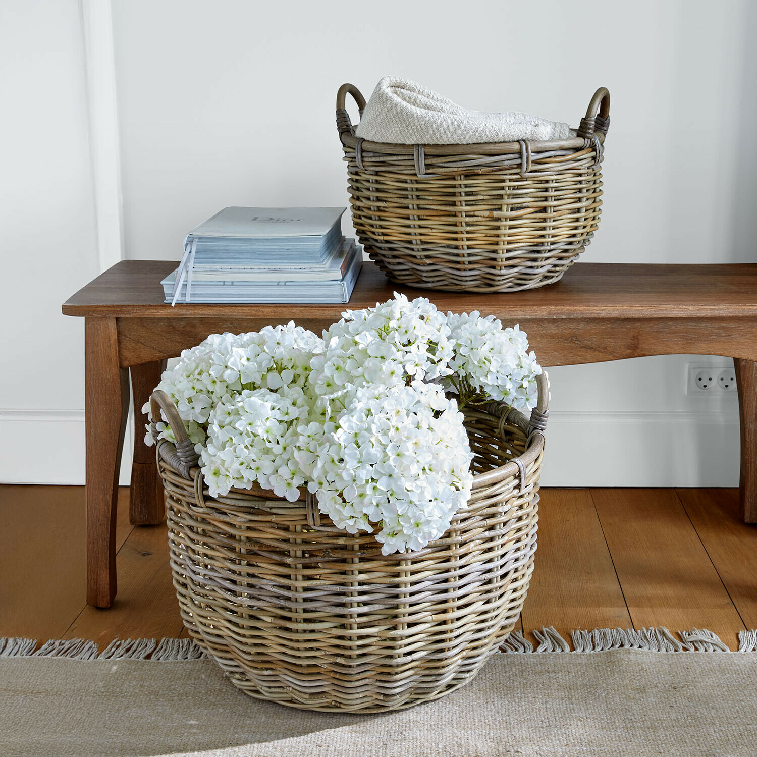 Grote rieten mand met witte bloemen en kleinere mand met deken op houten bank in woonkamer