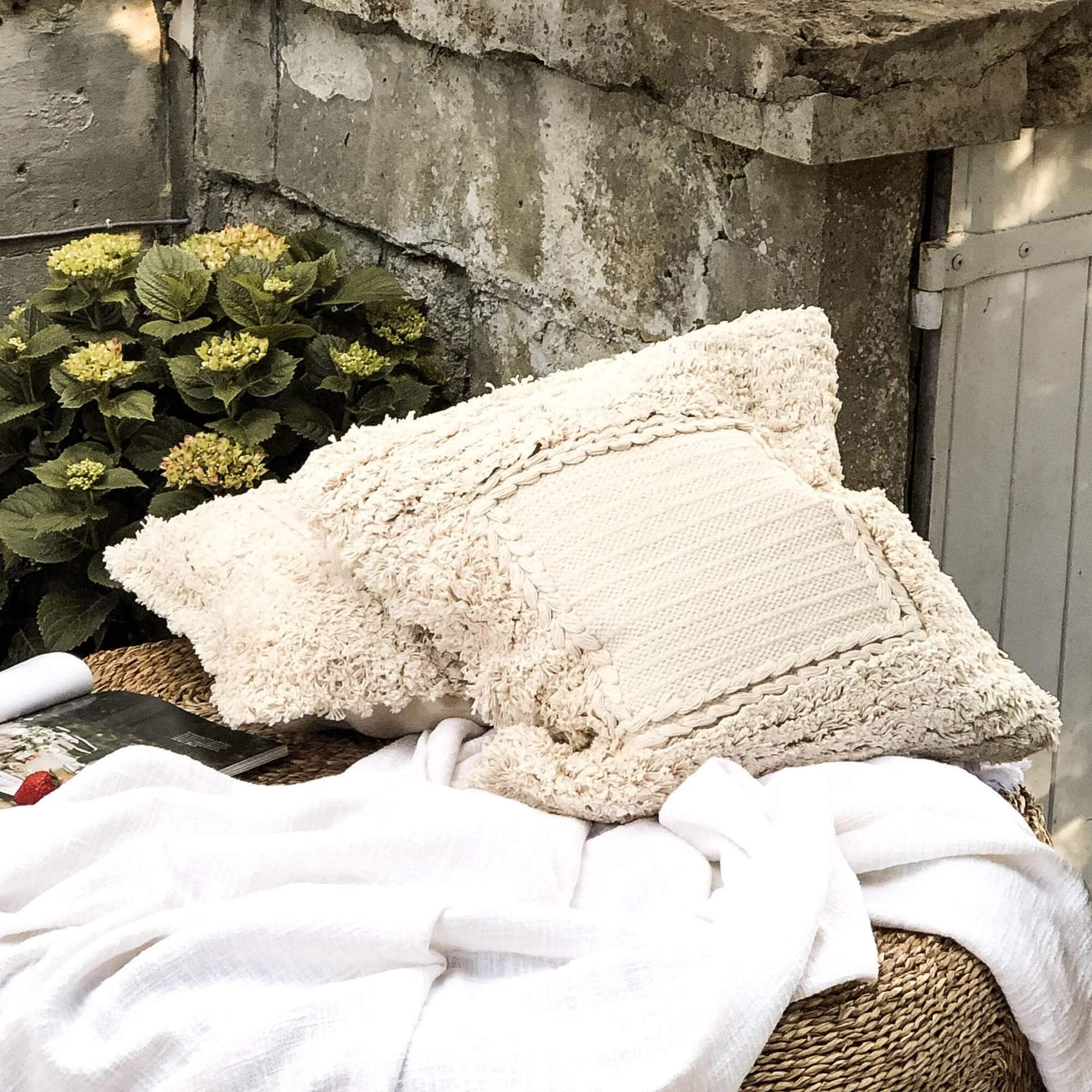 Beige decoratief kussen van zacht materiaal op gevlochten tuinmeubel met witte deken