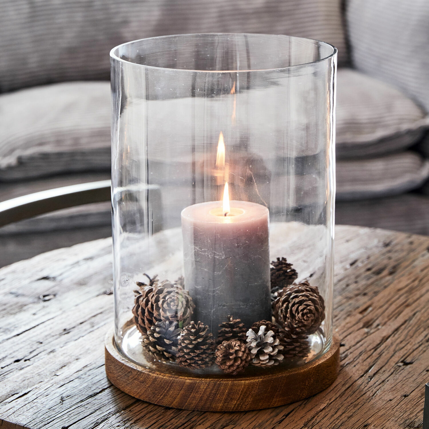 Grijze kaars in glazen windlicht met houten voet en dennenappels op rustieke tafel