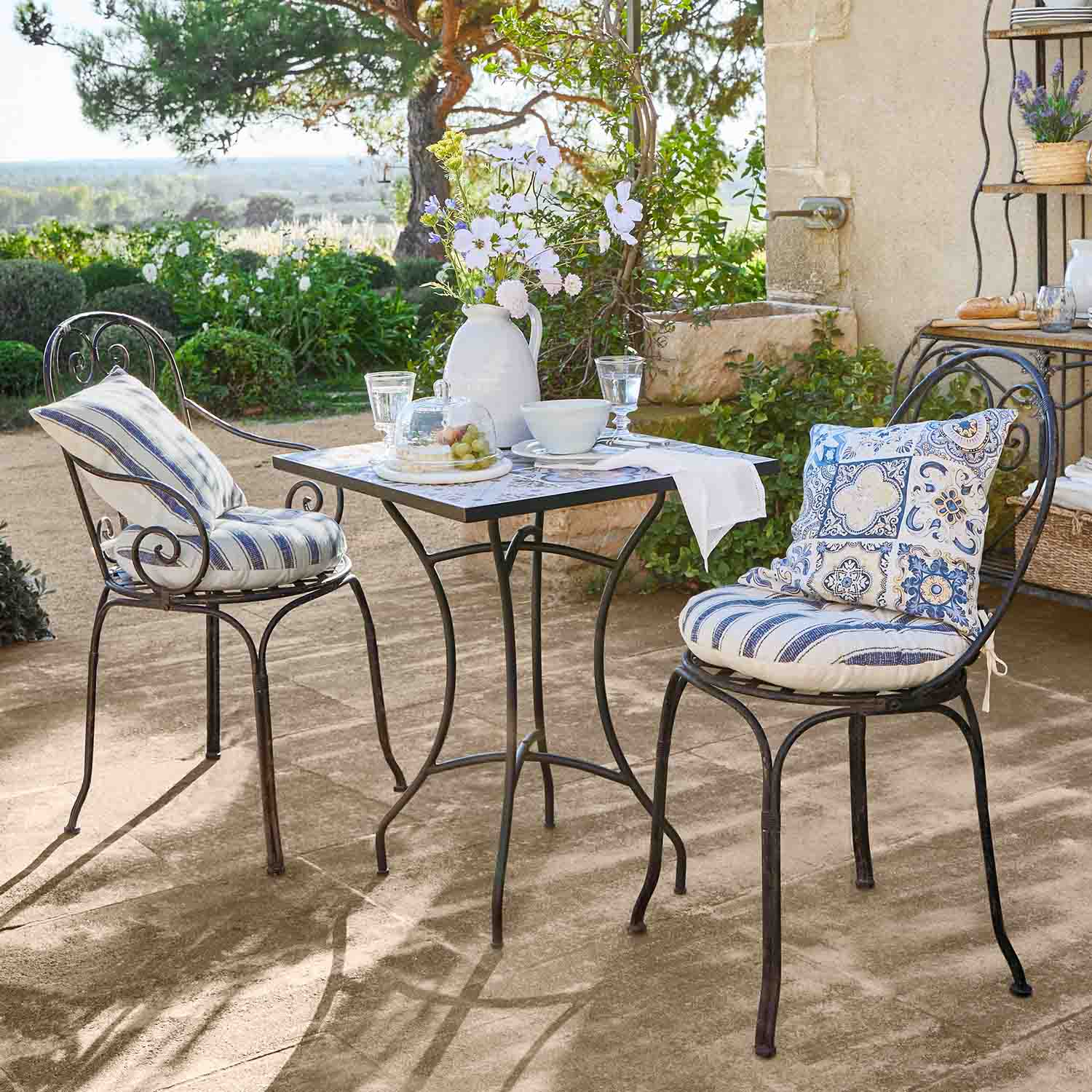 Metalen vierkante tafel met twee stoelen met blauwe kussens op zonnig tuinterras