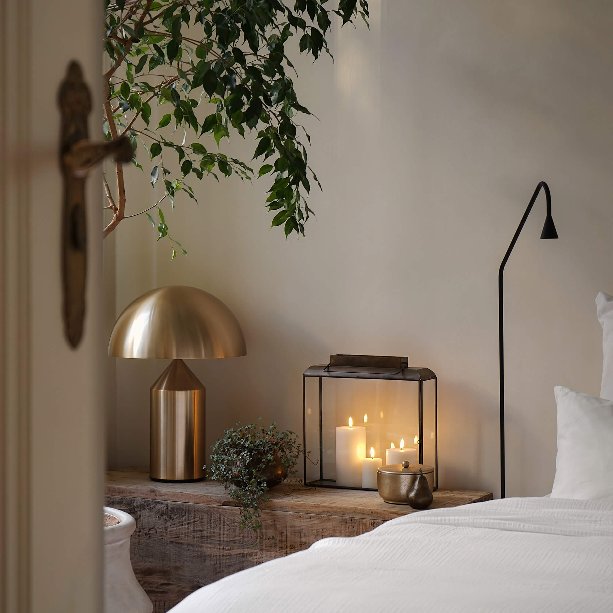 Gouden metalen tafellamp en glazen lantaarn met witte kaarsen op houten plank in slaapkamer