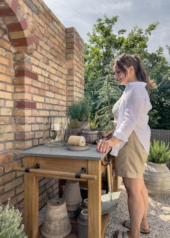 Vrouw in wit overhemd en beige korte broek bij houten tuintafel met betonnen blad voor een bakstenen muur en planten