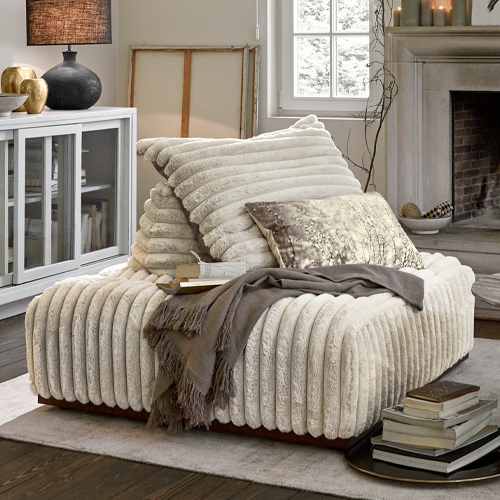 Beige fauteuil van zacht materiaal met kussens en grijze deken in gezellige woonkamer met open haard