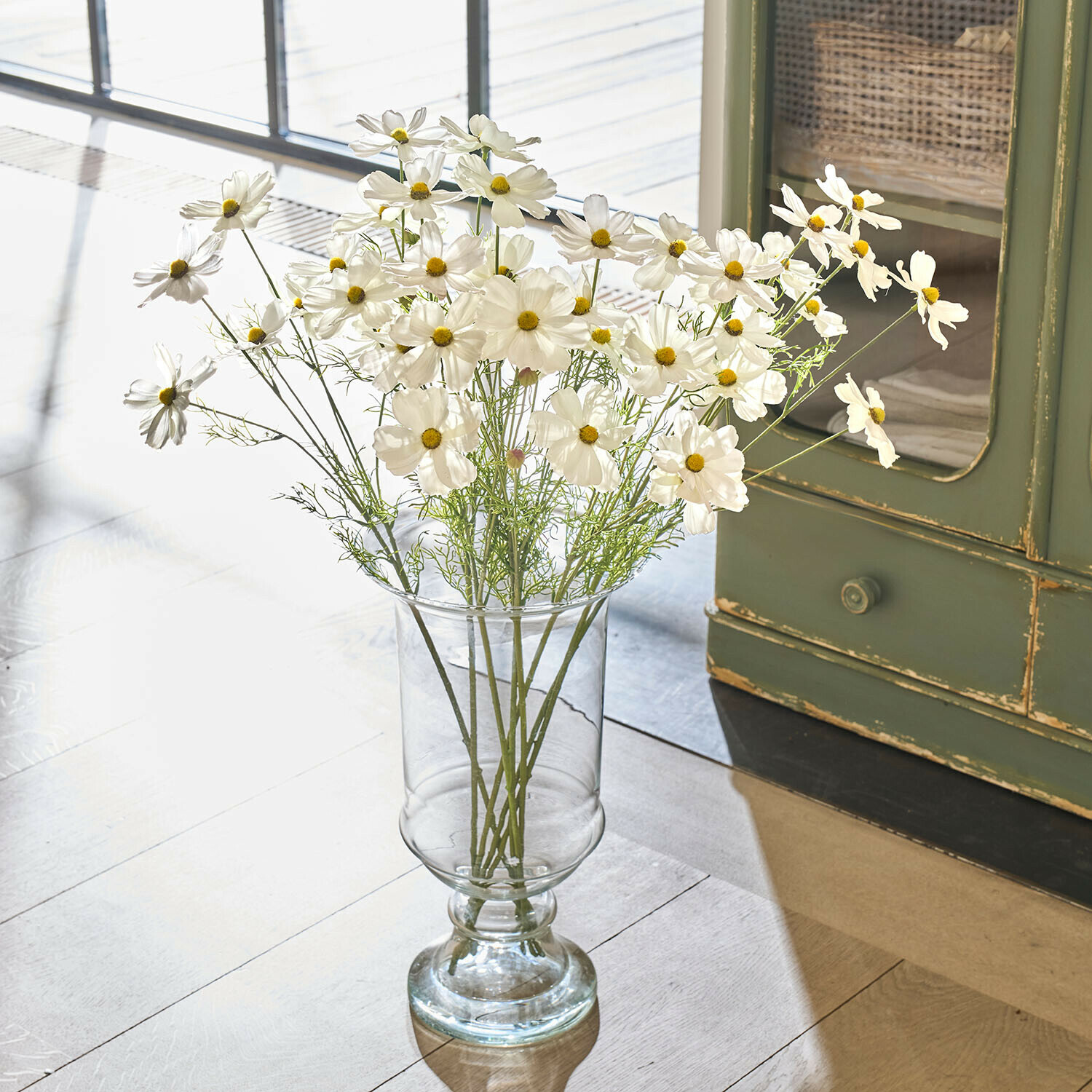 Transparante glazen vaas met witte kosmosbloemen op lichte vloer in natuurlijk licht