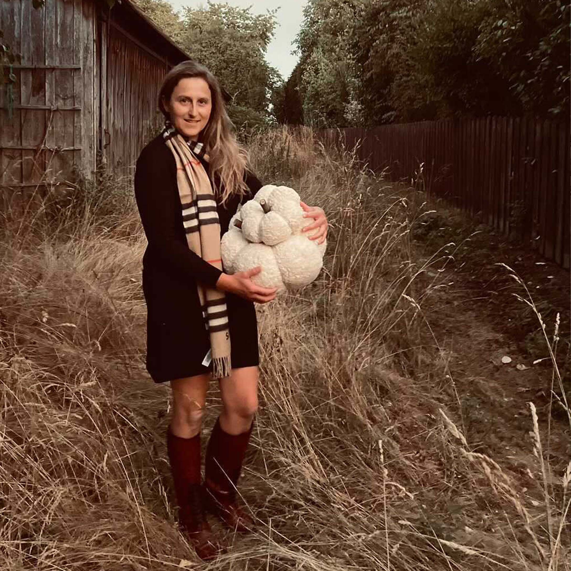 Vrouw in zwarte jurk en sjaal houdt witte decoratieve pompoen vast op een herfstveld