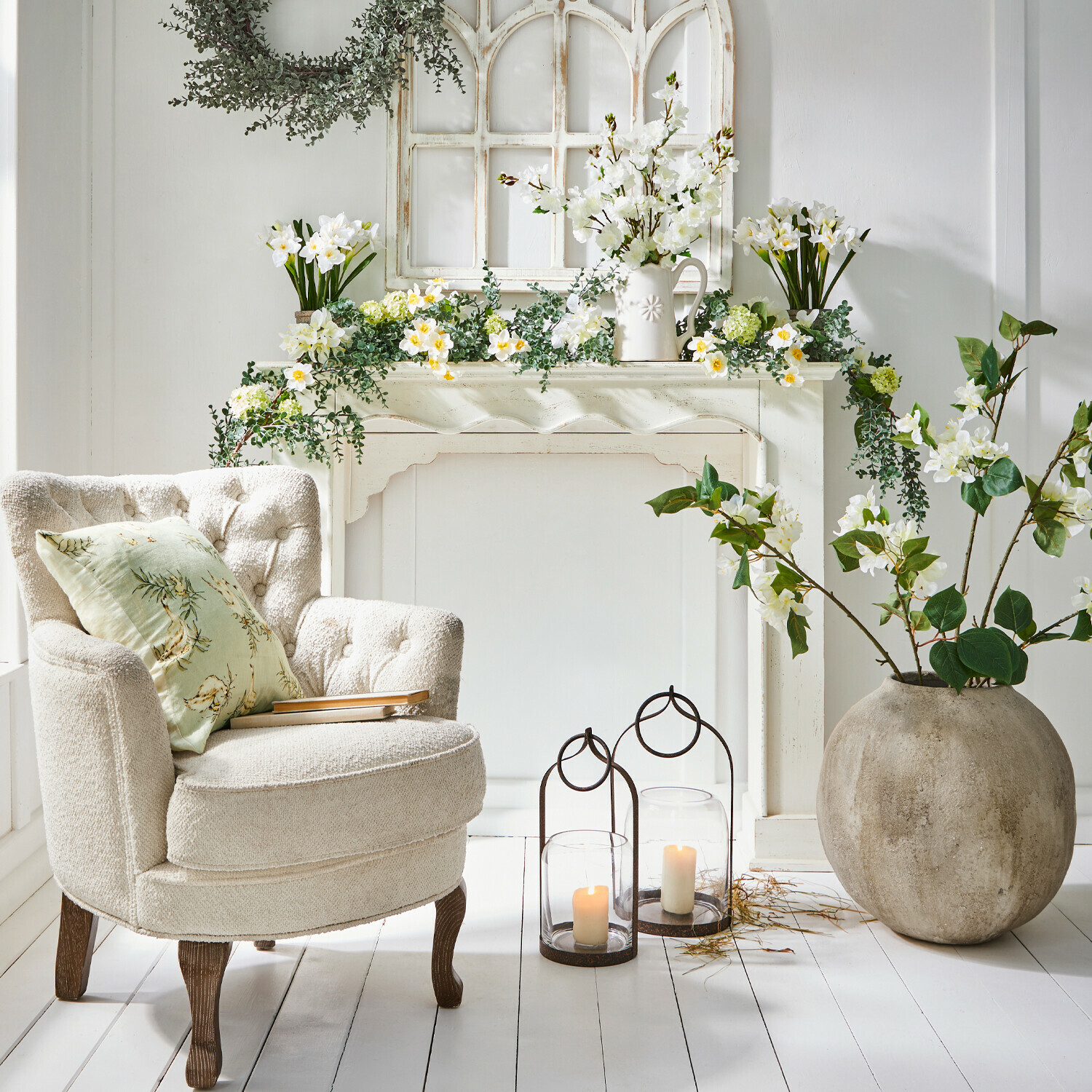 Haardschouw Claybourne Comfortabele crèmekleurige fauteuil met houten poten naast decoratieve schouw met witte bloemen en kaarsen