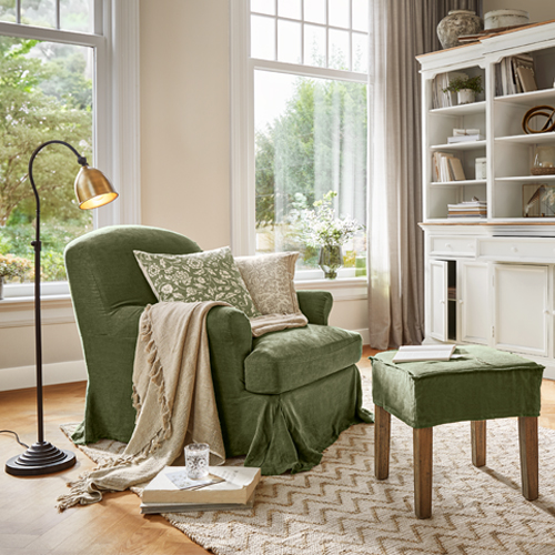 Groene stoffen fauteuil met voetenbank en beige deken in lichte woonkamer met groot raam