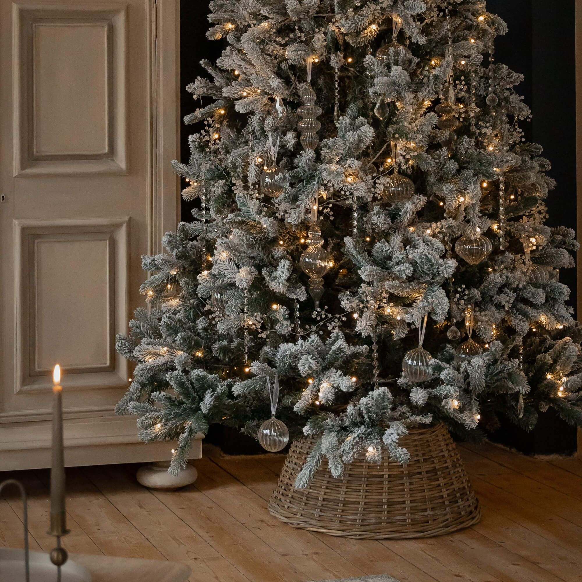 Besneeuwde kerstboom met glazen versieringen en warme lichtjes op een rieten standaard