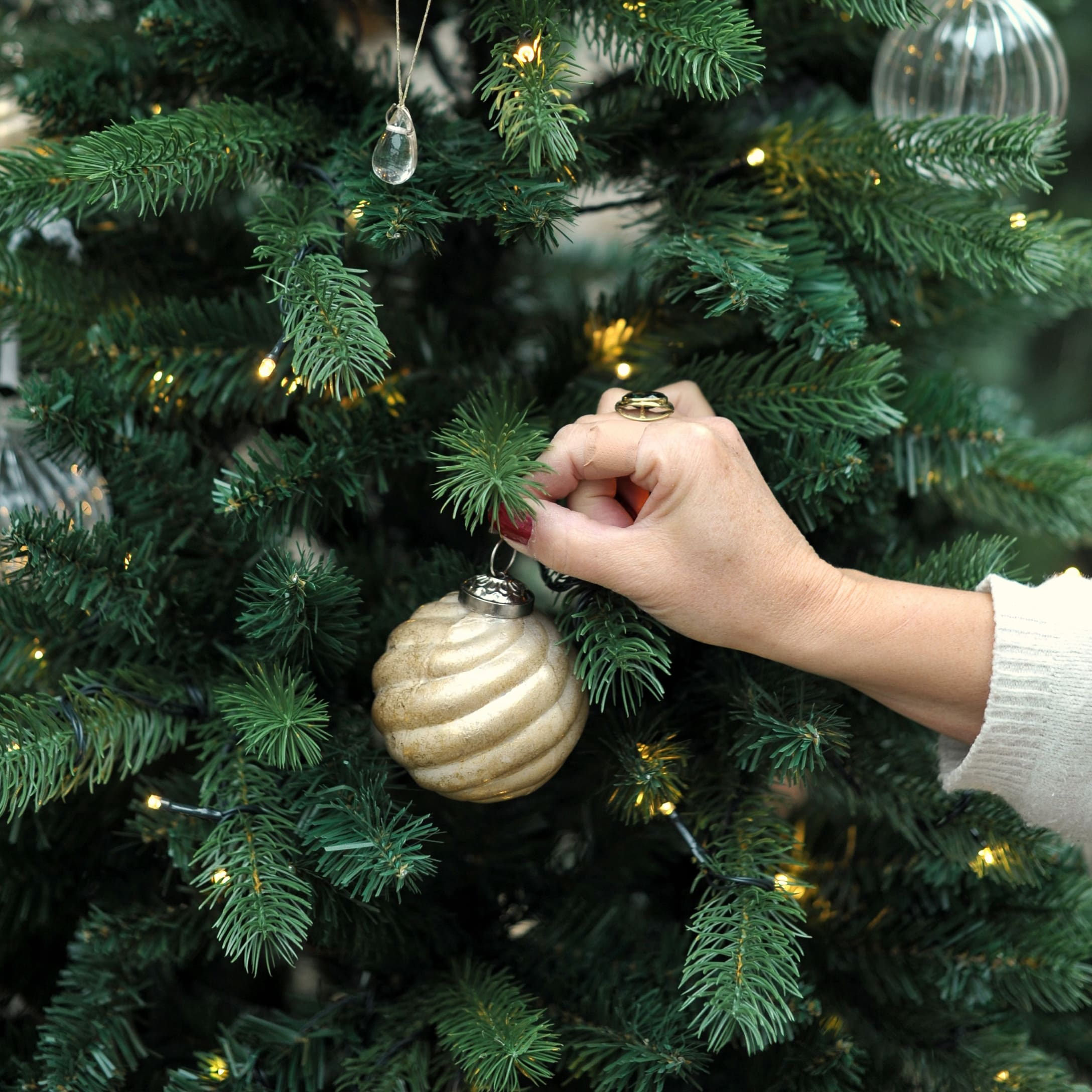Hand hangt gouden kerstbal aan groene kerstboom met LED-lichtjes in serre