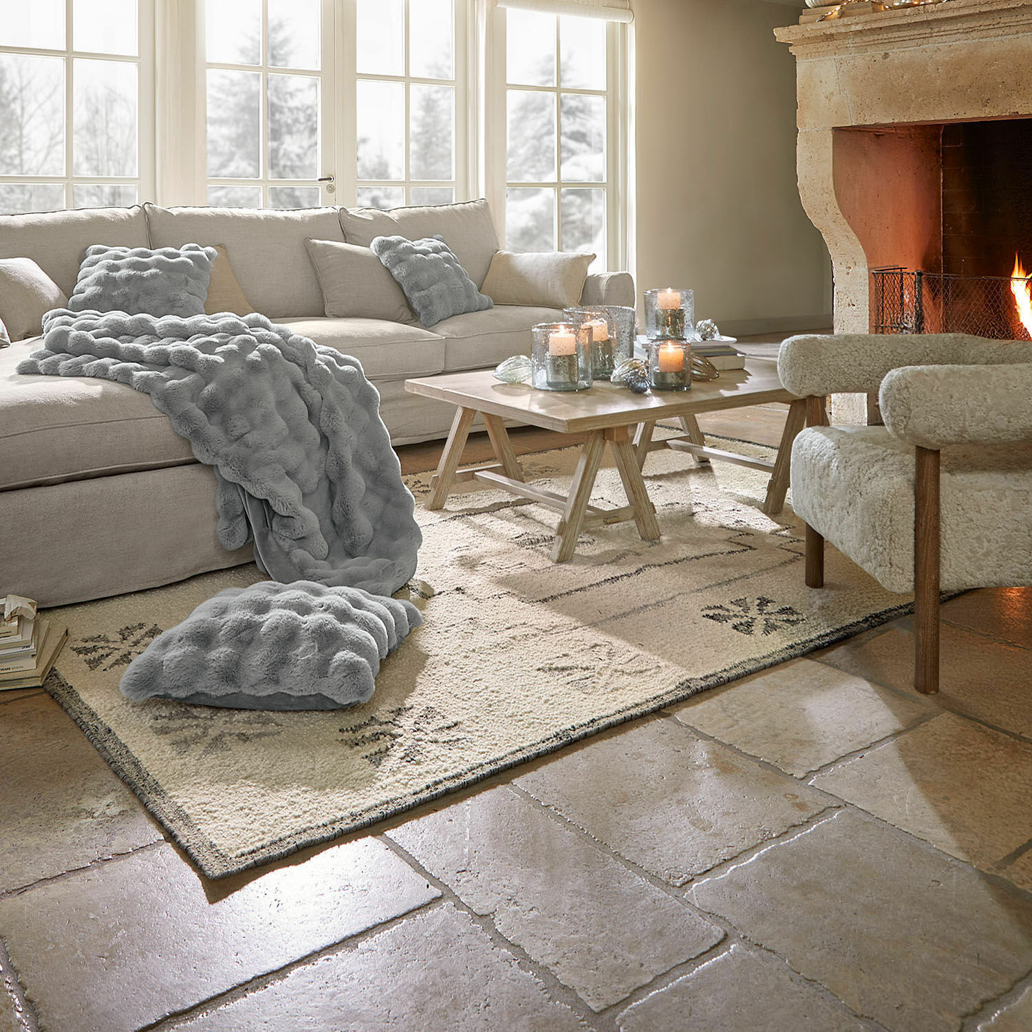 Gezellige woonkamer met beige bank, grijze deken en kussens, brandende open haard en houten tafel