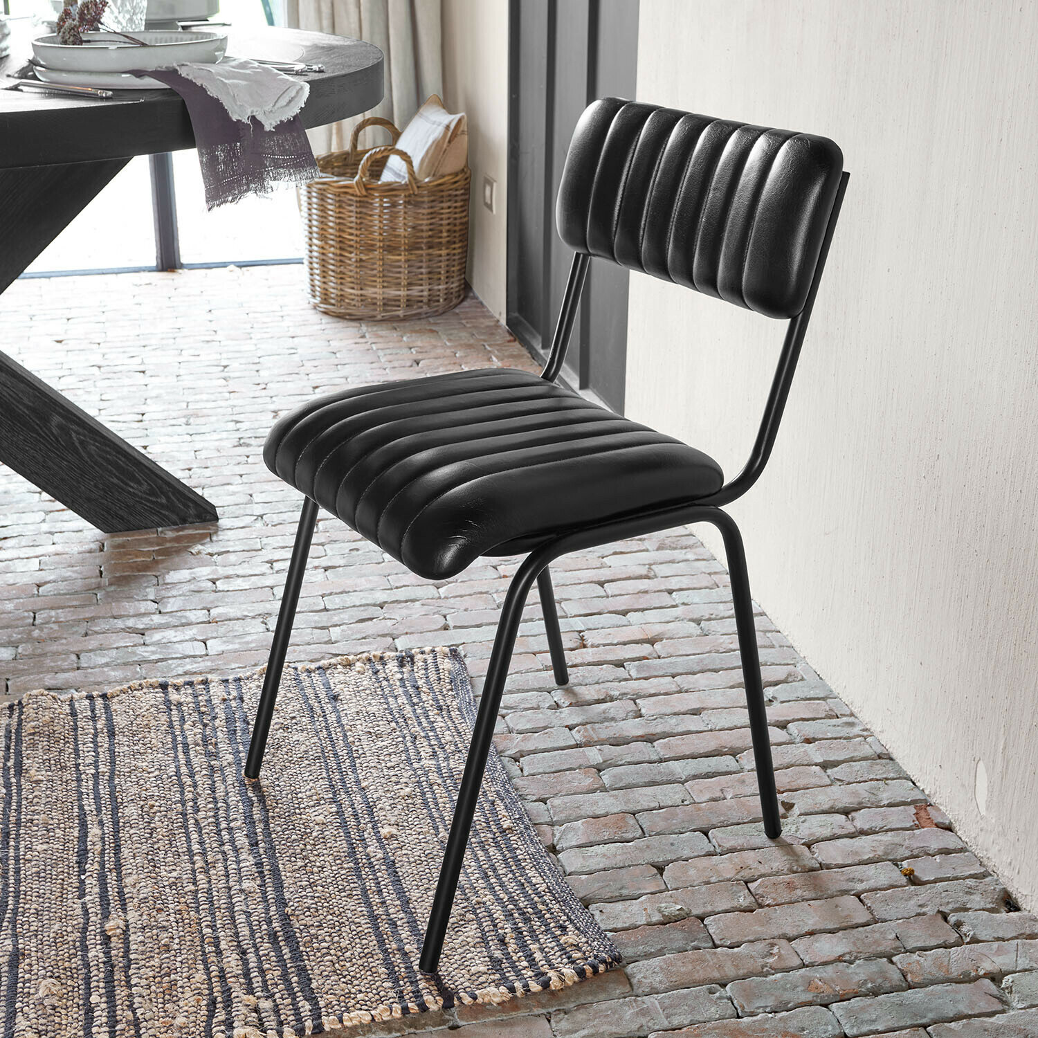 Stoel Jolivon Zwarte stoel met doorgestikte leren bekleding en metalen frame op bakstenen vloer naast vloerkleed