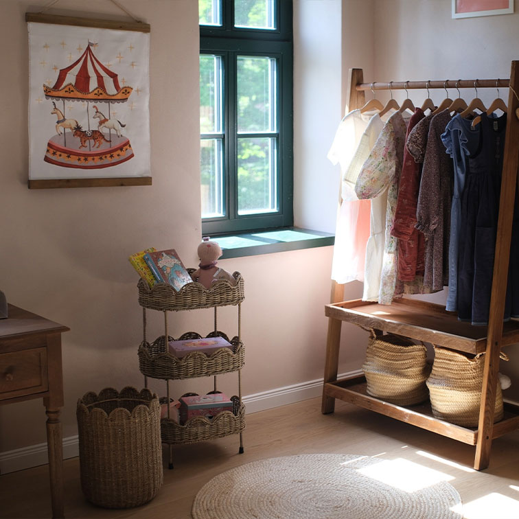 Drie verdiepingen rieten rek met speelgoed en kinderkleding aan houten kledingrek in lichte kamer