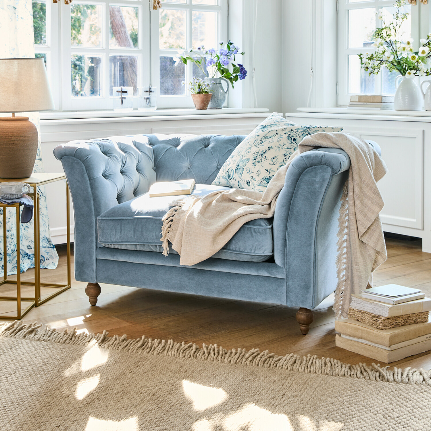 Blauwe fluwelen tweezitsbank met gestikte rugleuning en beige deken in lichte woonkamer