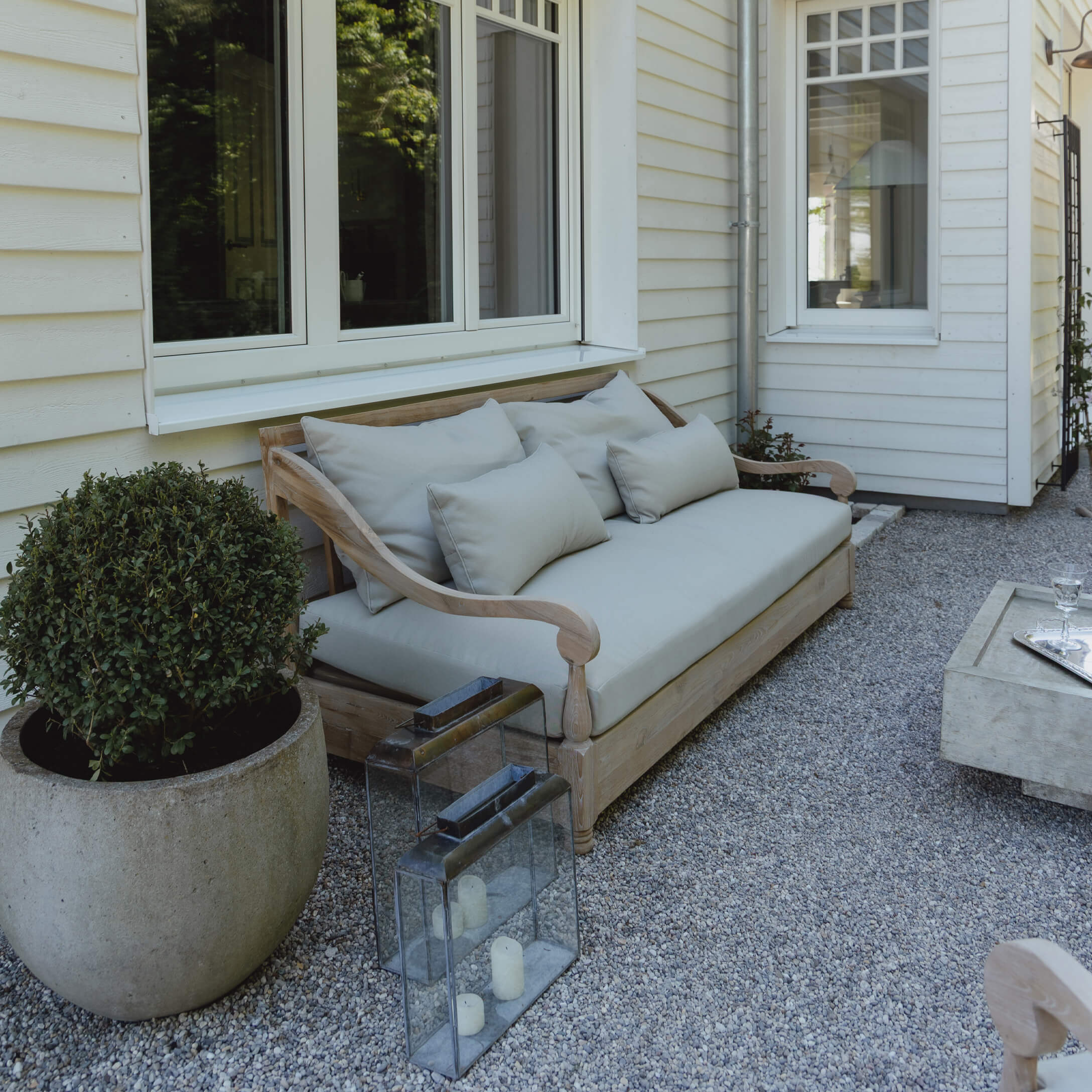 Houten loungesofa met lichtgrijze kussens op grindpatio bij wit huis