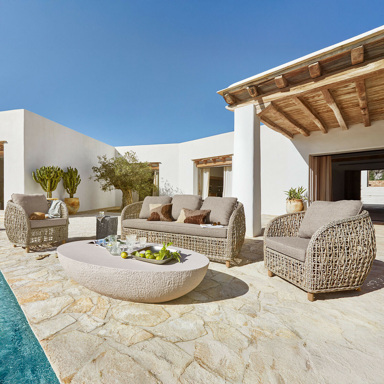 Beige rotan tuinmeubelset met kussens op stenen terras bij zwembad in zonnige buitenruimte