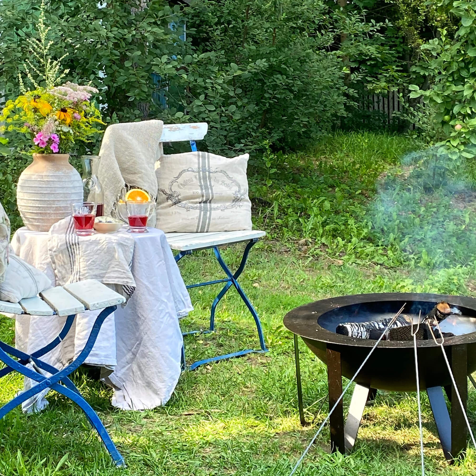 Tuin met blauwe metalen stoelen, wit tafelkleed en zwarte vuurkorf op groen gras