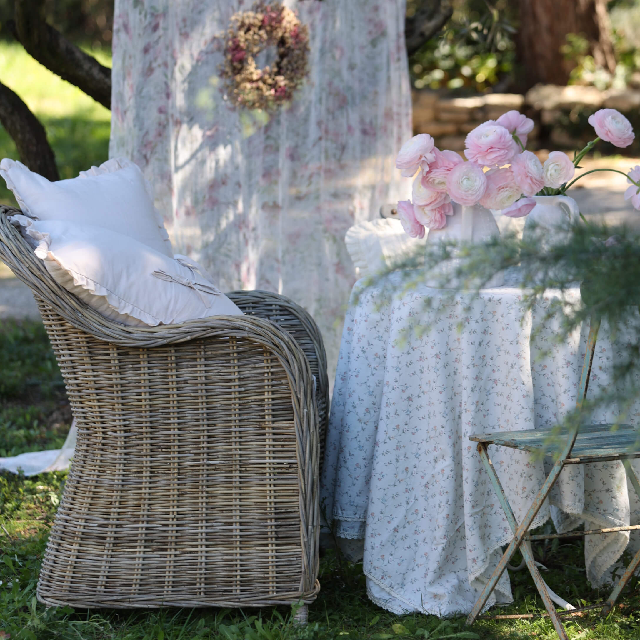 Rotan stoel met witte kussens naast tafel met bloemen tafelkleed en roze bloemen in tuin