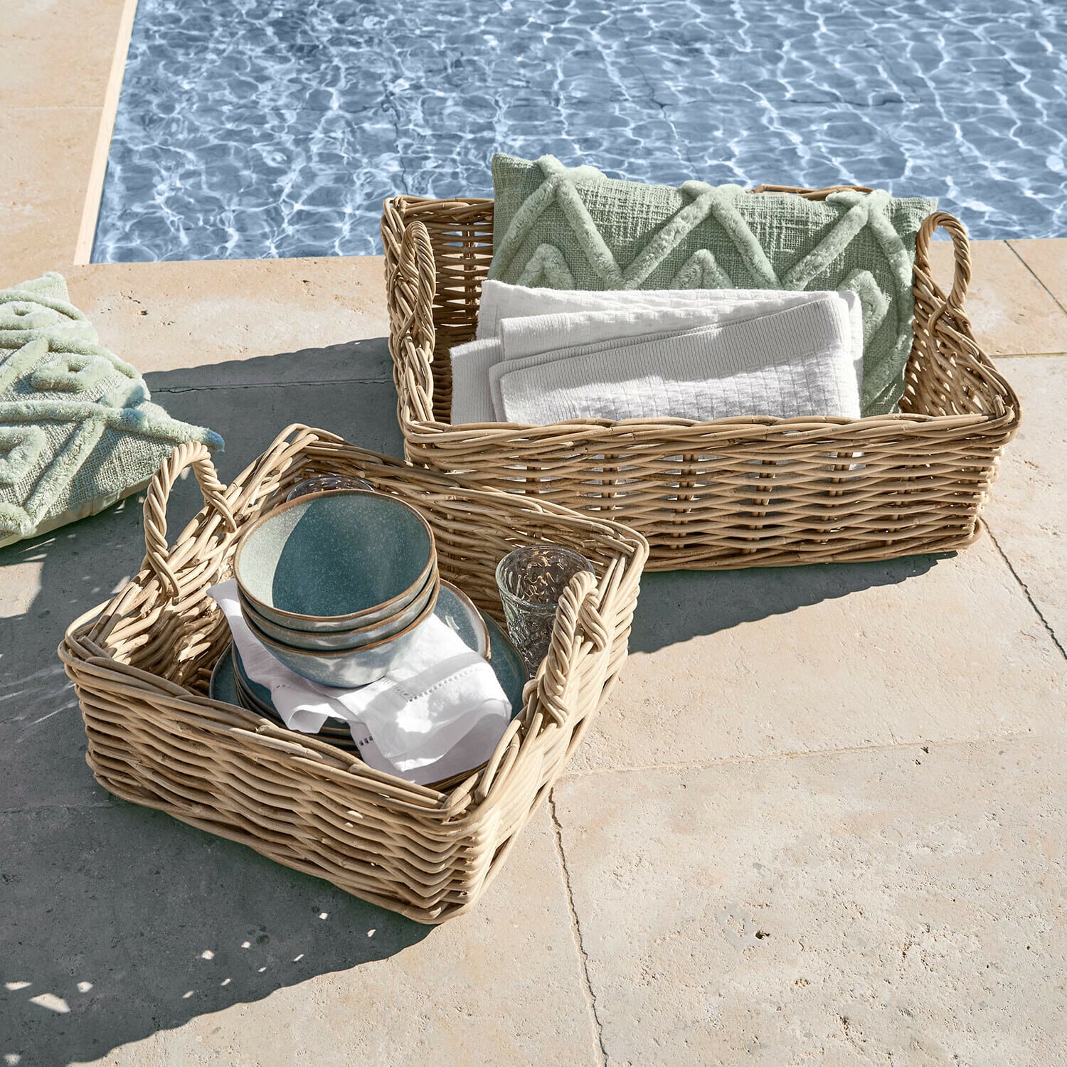 Twee rieten manden met handvatten, gevuld met groene kussens en witte handdoeken bij het zwembad