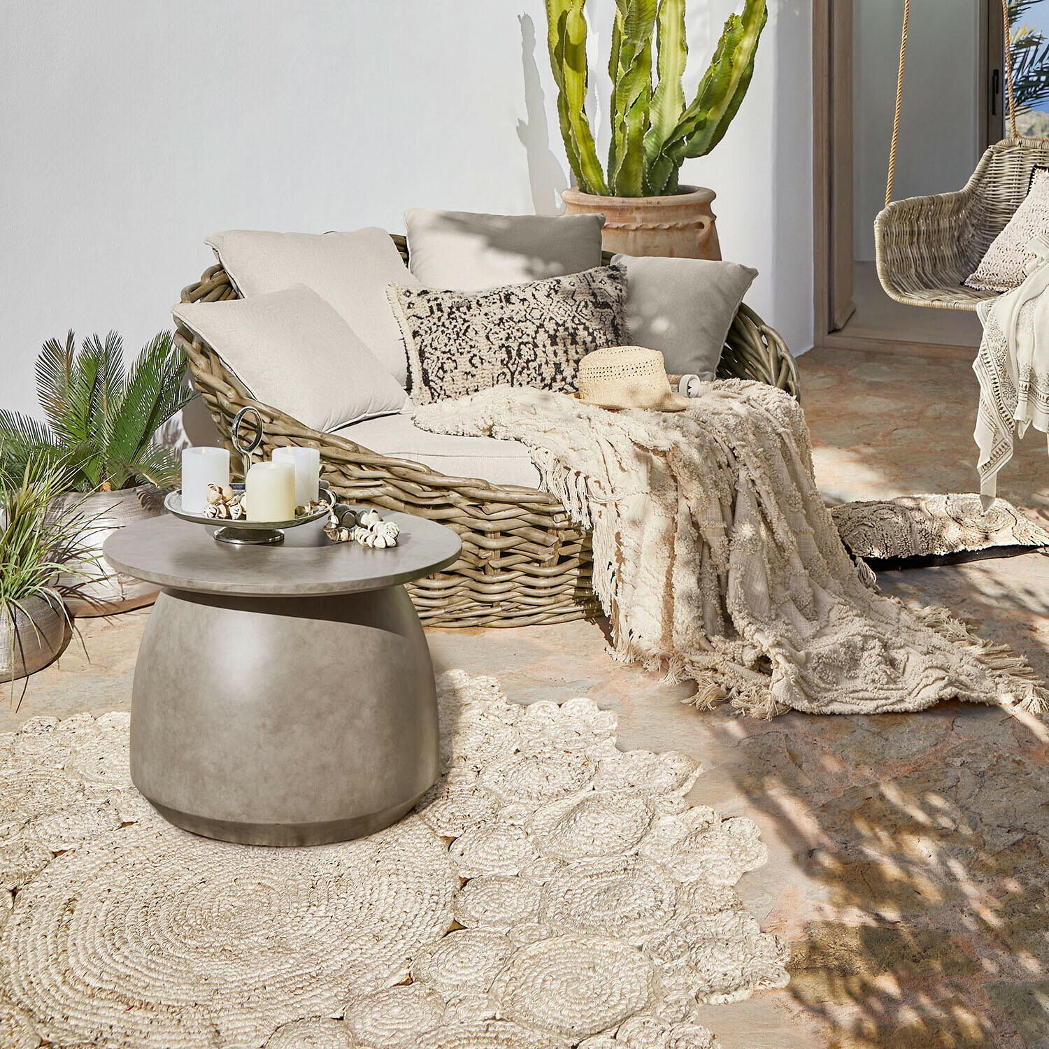 Ronde betonnen salontafel op terras met rotan bank en crèmekleurige decoratieve deken