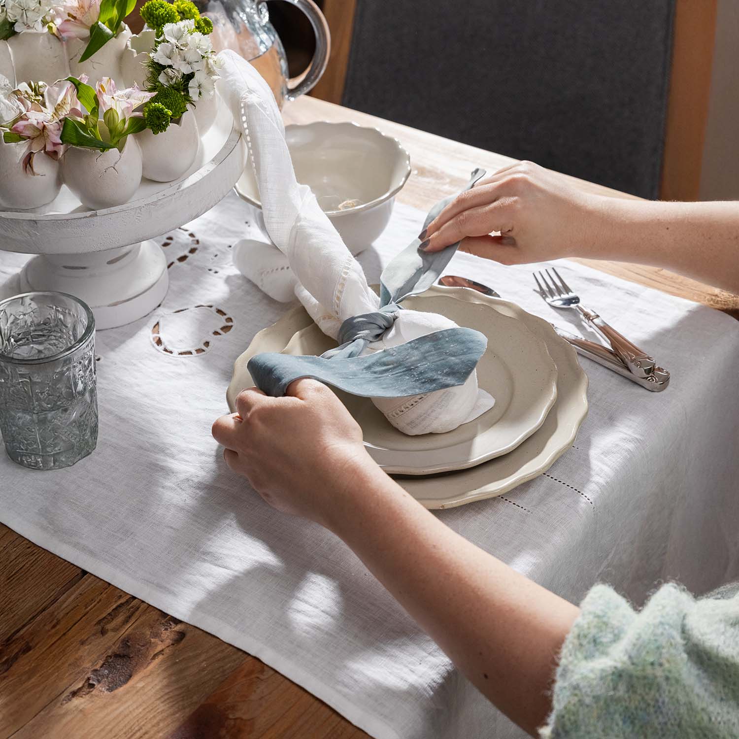 Handen die een wit linnen tafelkleed en servet met blauwe strik op een houten tafel met porseleinen borden leggen