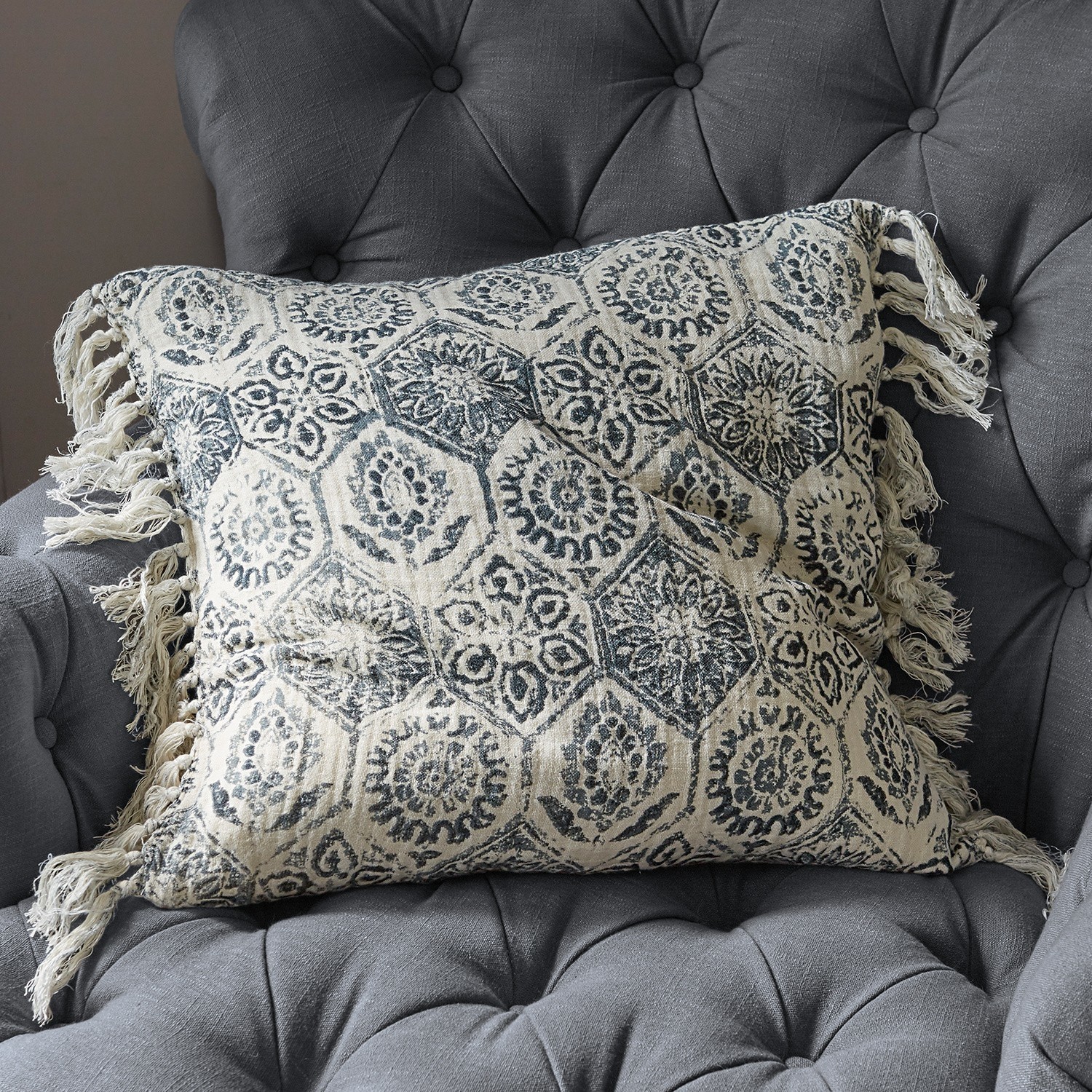 Beige katoenen kussen met blauw patroon en franjes op een grijze gestoffeerde stoel